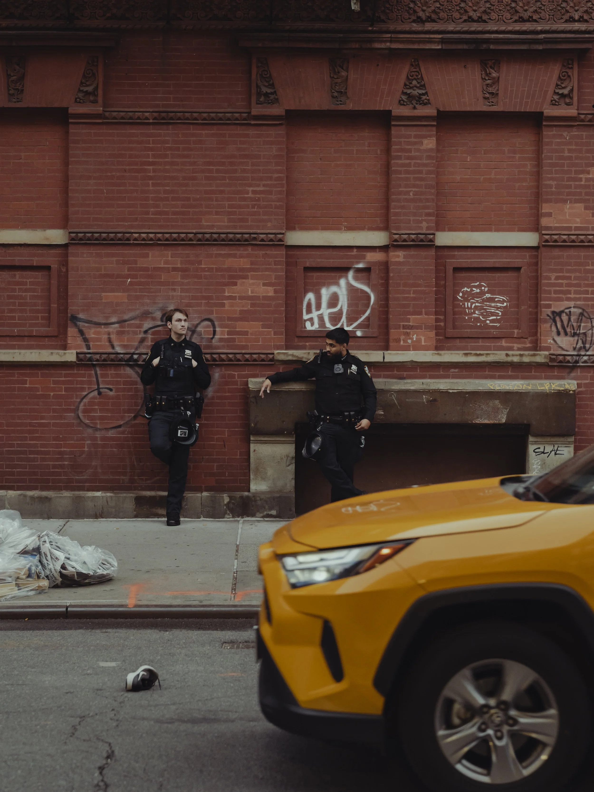 Two police officers leaning against a red brick wall with graffiti, a yellow car partially visible in the foreground, and trash bags on the sidewalk.