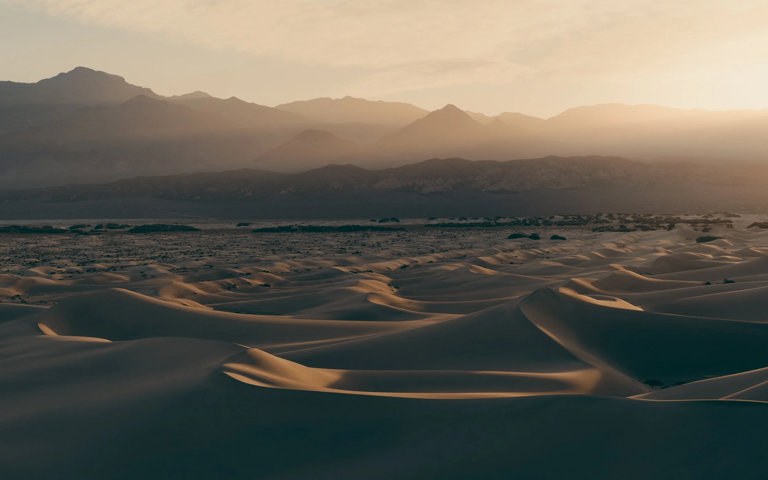 Sand dunes in a desert with mountains in the background and the sun setting.