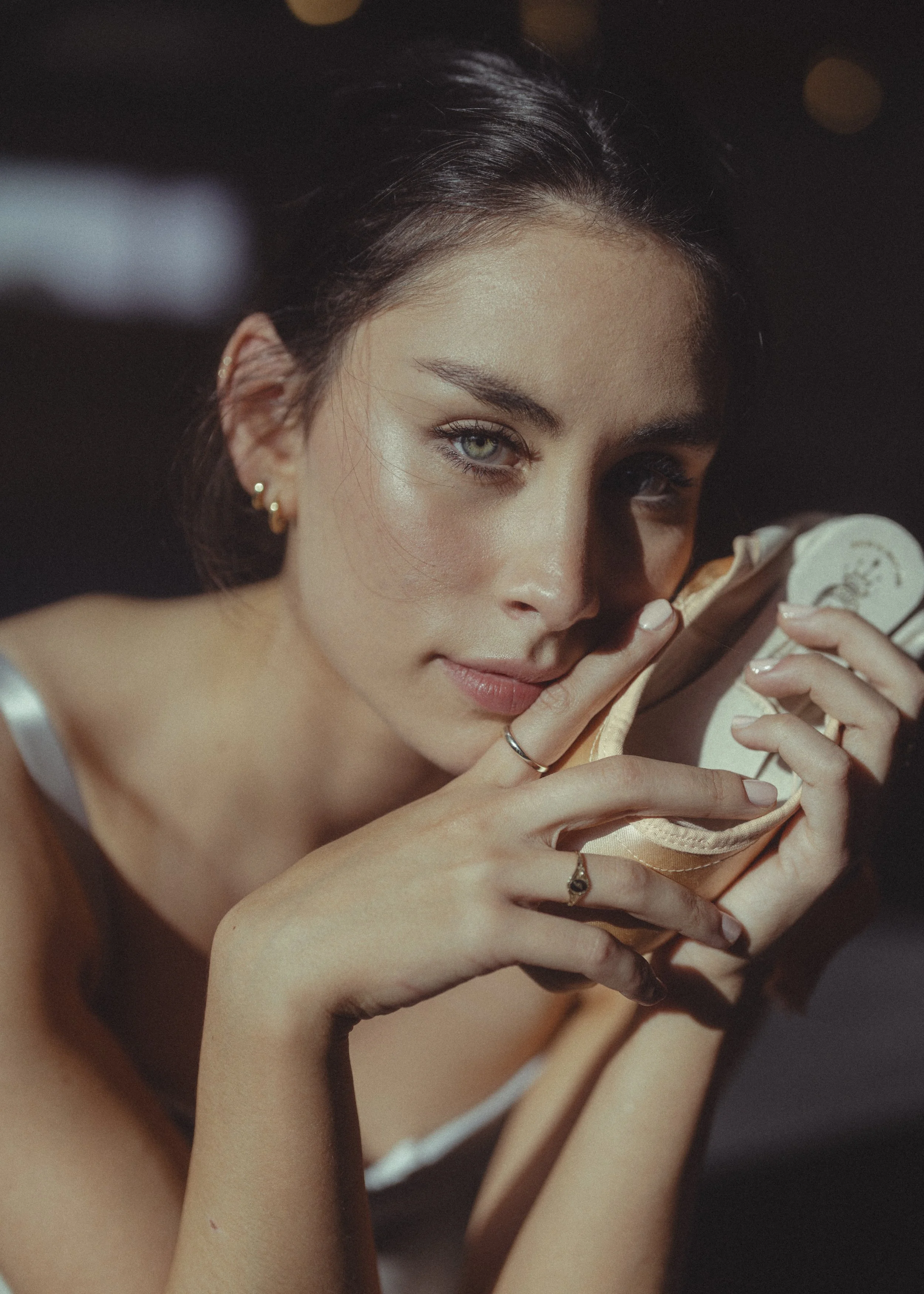 A young woman with dark hair wearing earrings holds a light-colored cloth to her face, looking at the camera with a soft expression.