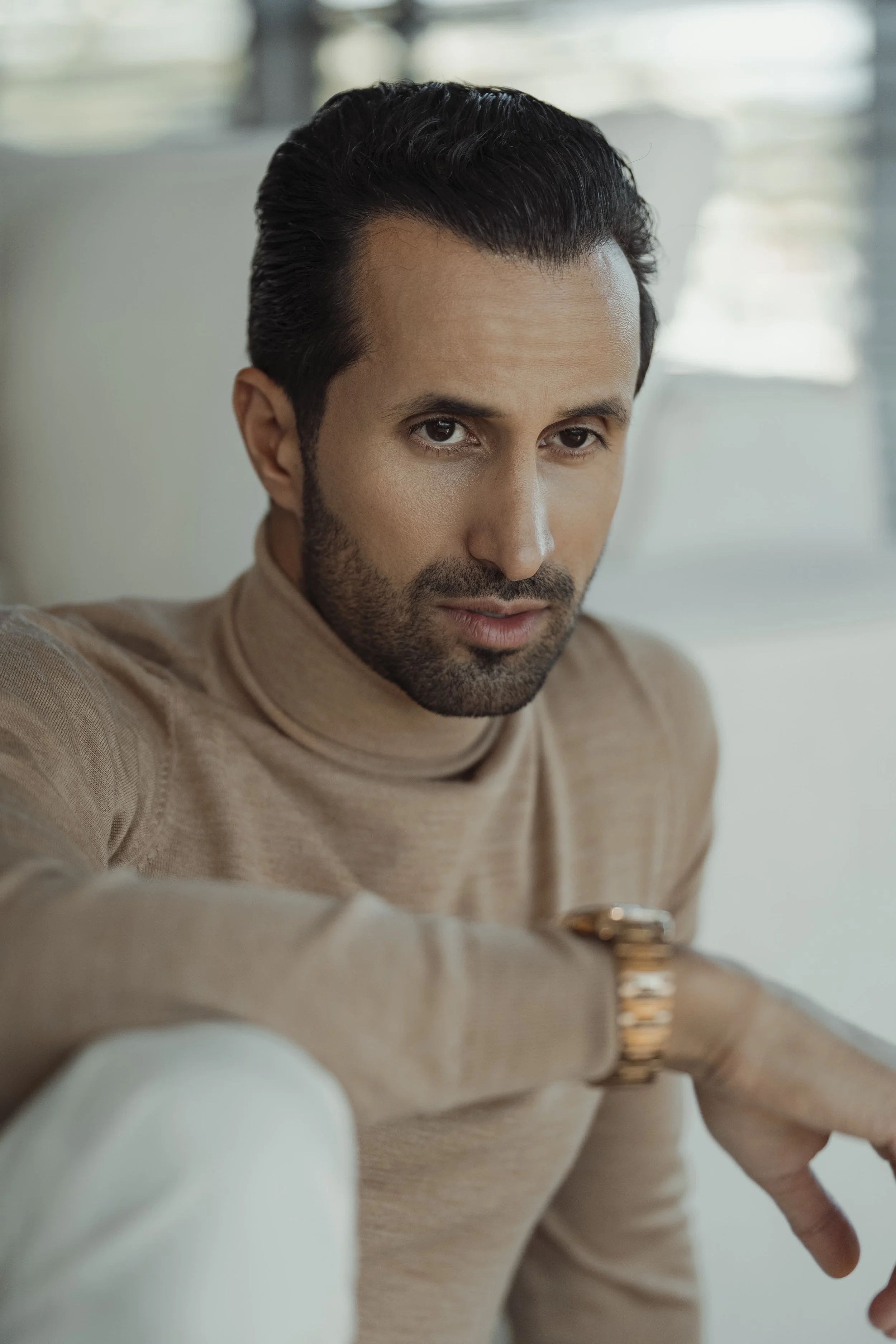A man with dark hair and a beard wearing a beige turtleneck sweater, looking at the camera with a serious expression.