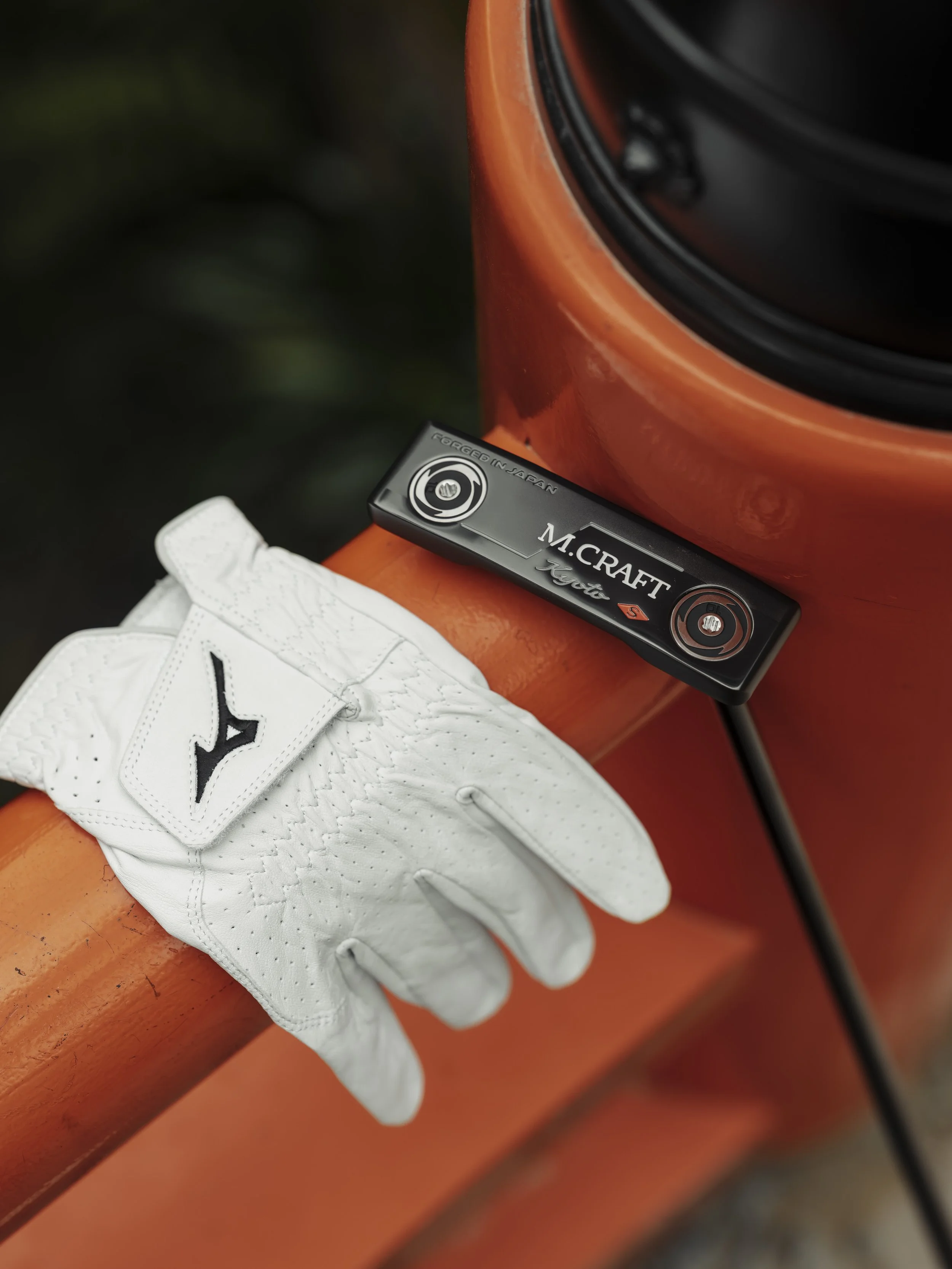 A white golf glove with a black Mizuno logo resting on an orange golf club with a M.CRAFT golf club headcover placed on top.