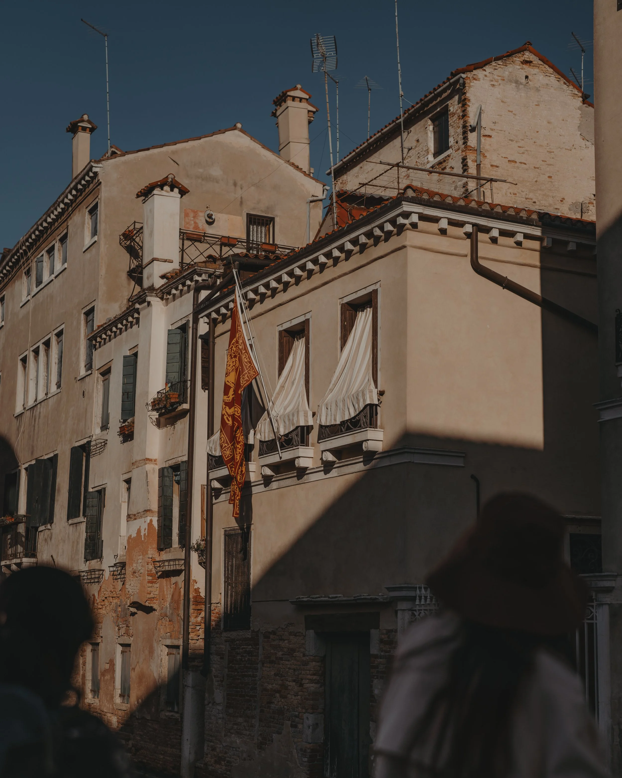 A cluster of old buildings with beige and white facades, some with peeling paint, with terracotta tile roofs and multiple antennas on top. There are two balconies with white awnings, a red flag hanging from one, and the scene is partly shaded by a sh