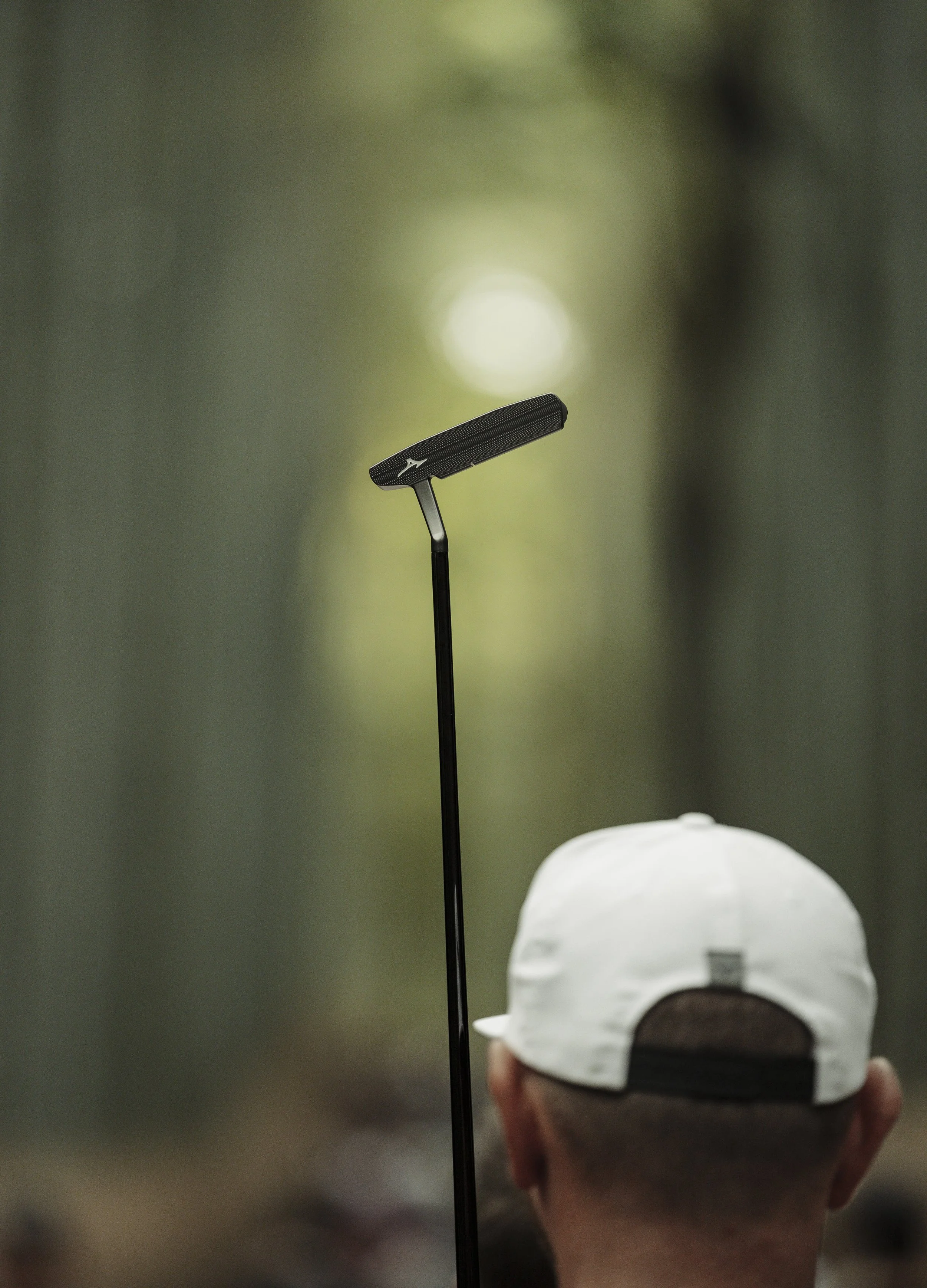 Close-up of a golf club held vertically behind a person wearing a white cap, with the person facing away from the camera in a wooded area.