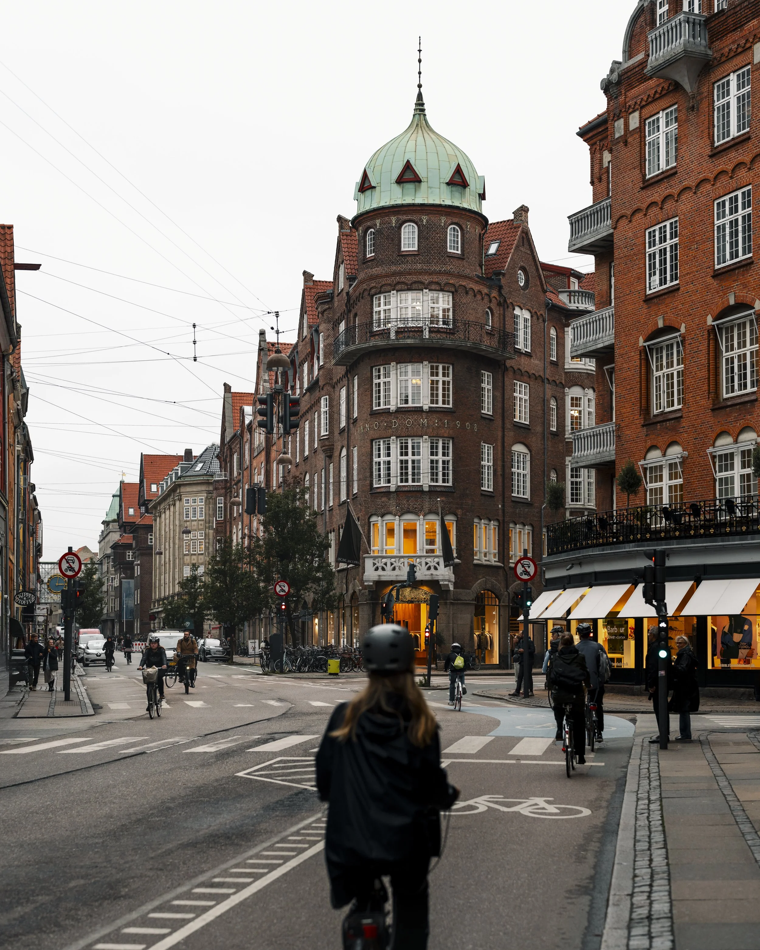 A street scene in a European city with cyclists and pedestrians, featuring historic brick buildings and a corner building with a green domed roof, during dusk with warm lights in the windows.