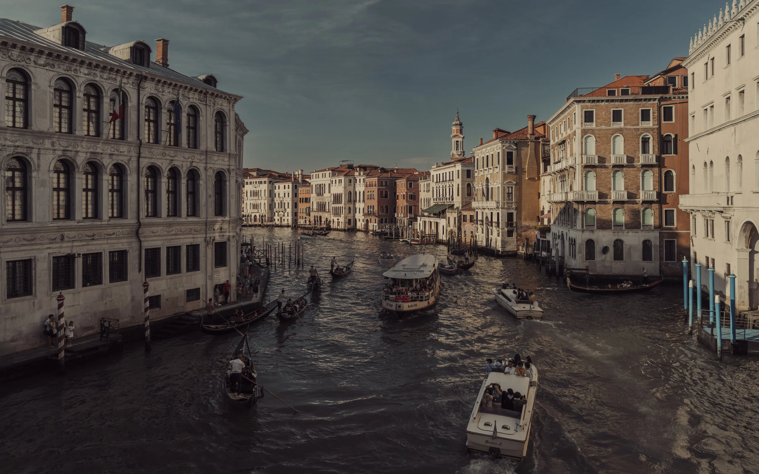 Gondolas and boats on a canal in Venice, Italy, with historic buildings lining the waterway under a partly cloudy sky.