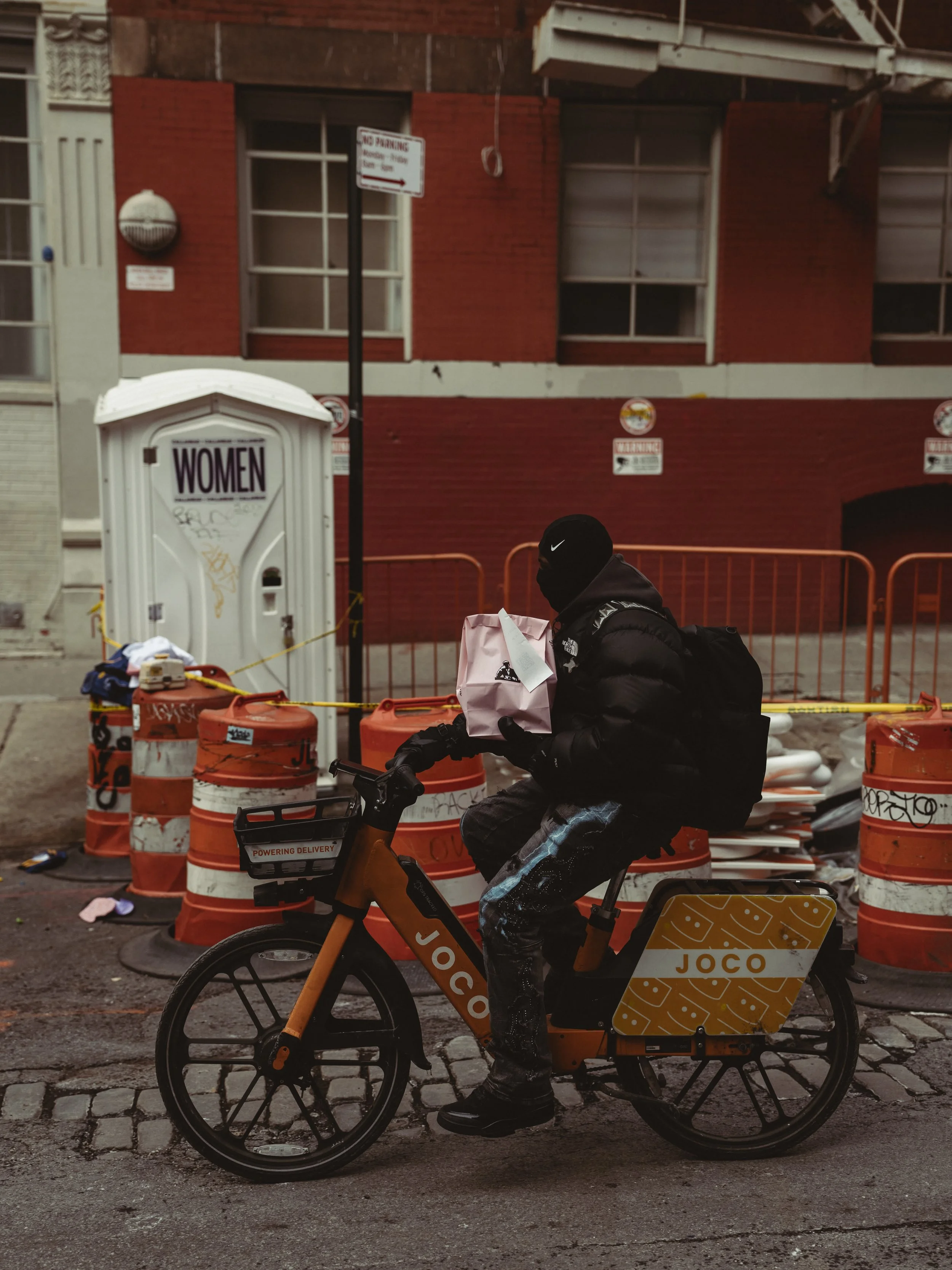 A delivery person on a Joco bike holding a pink paper bag on a city street, with orange and white construction barrels, a portable toilet, and a brick building in the background.