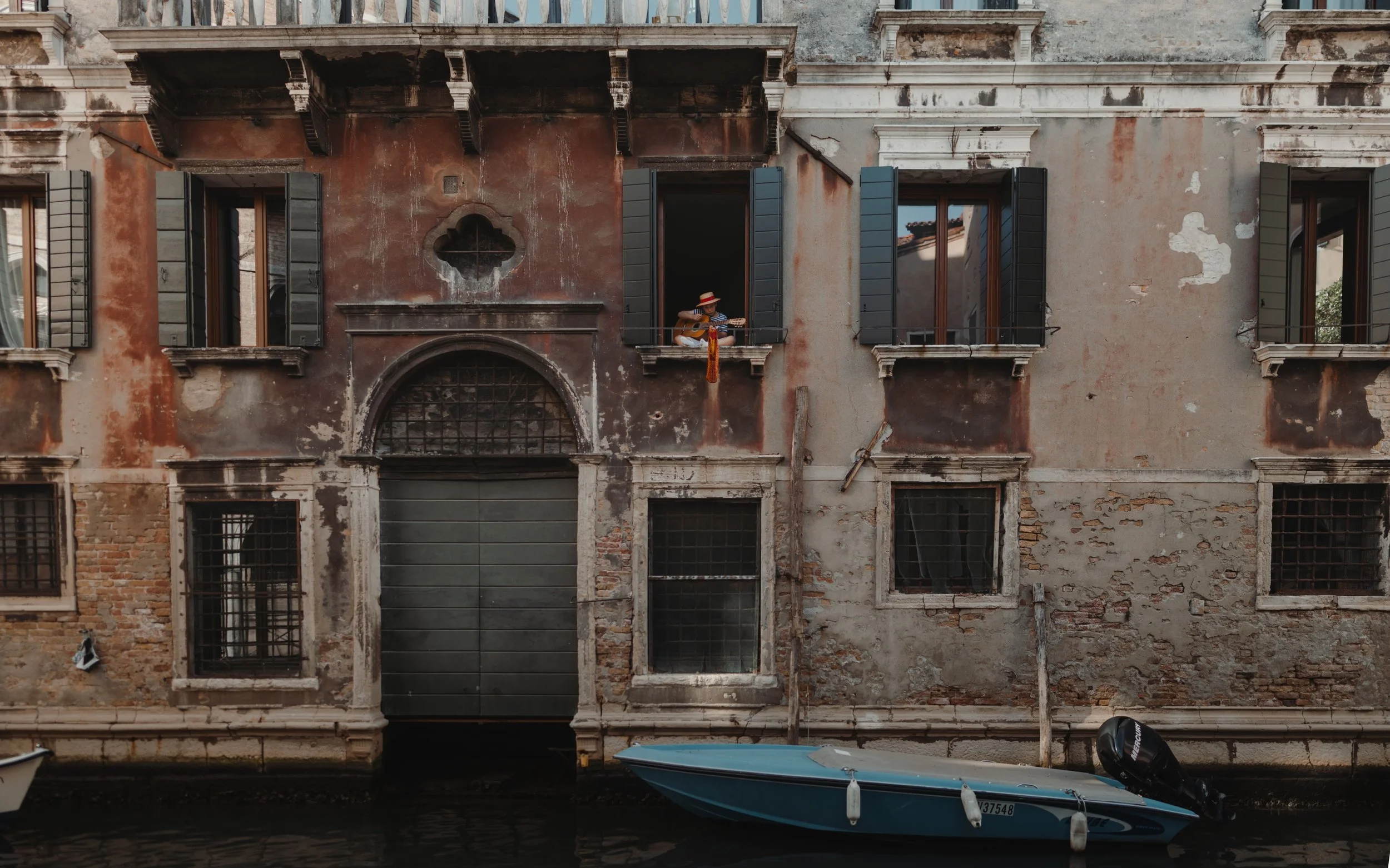 A woman sitting on a balcony in Venice, Italy, playing a guitar and wearing a striped hat with flowers, overlooking a canal with boats and an aged building with open windows and shutters.
