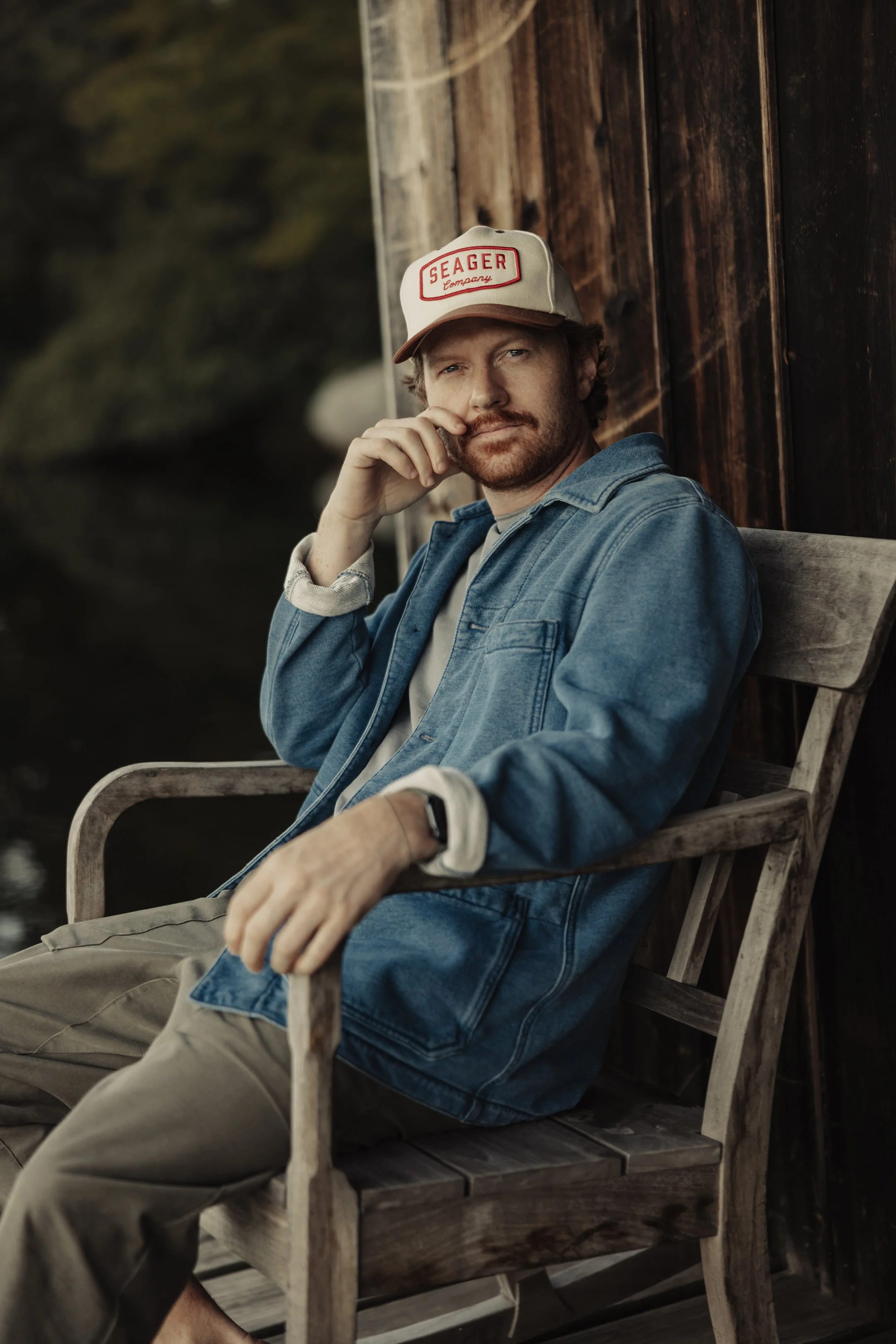 A man sitting on a wooden bench outdoors, wearing a denim jacket and a beige cap with a red logo, looking at the camera with a thoughtful expression.