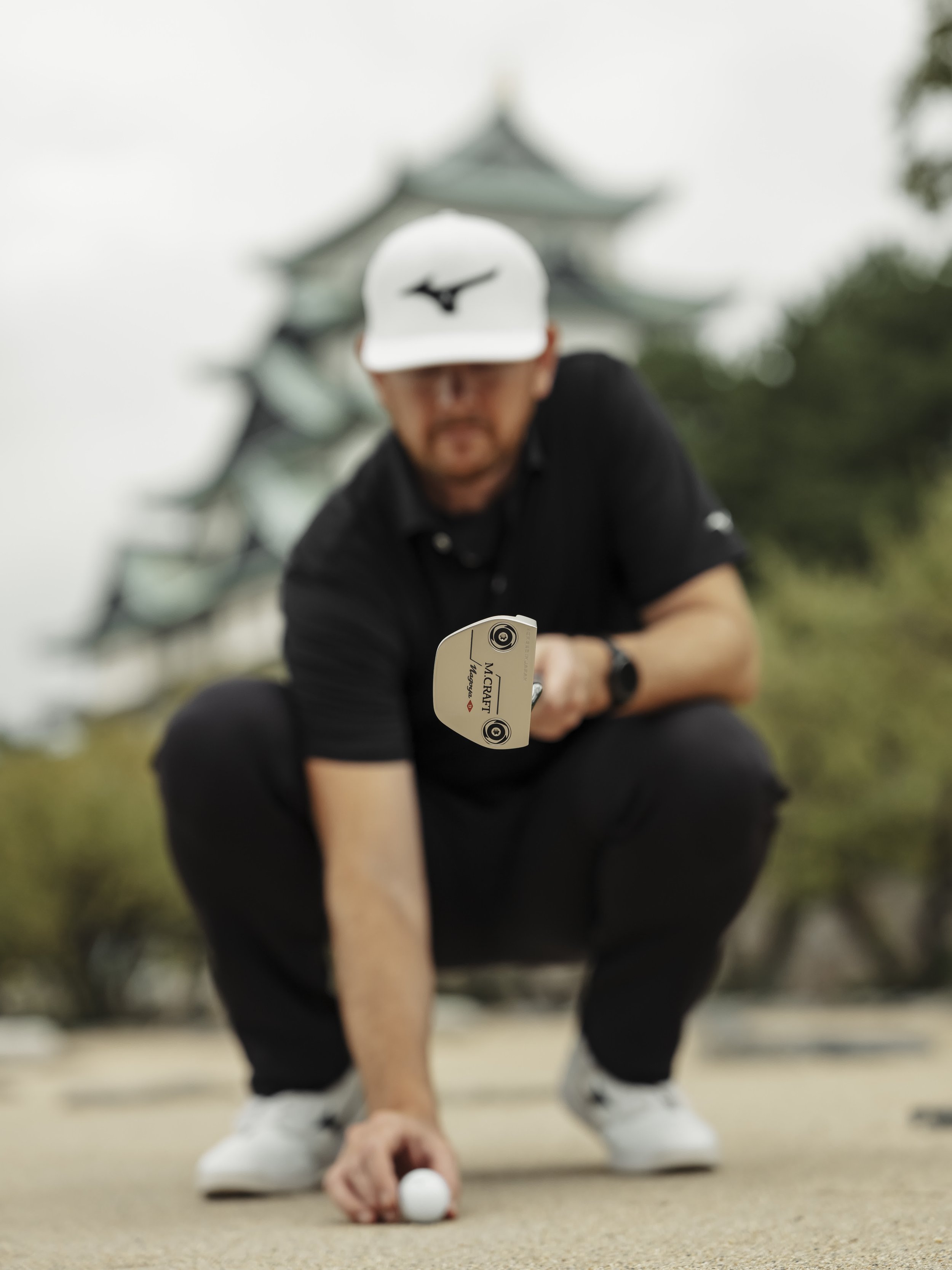A man crouching on a golf course preparing to hit a golf ball, with a traditional Japanese castle in the background.