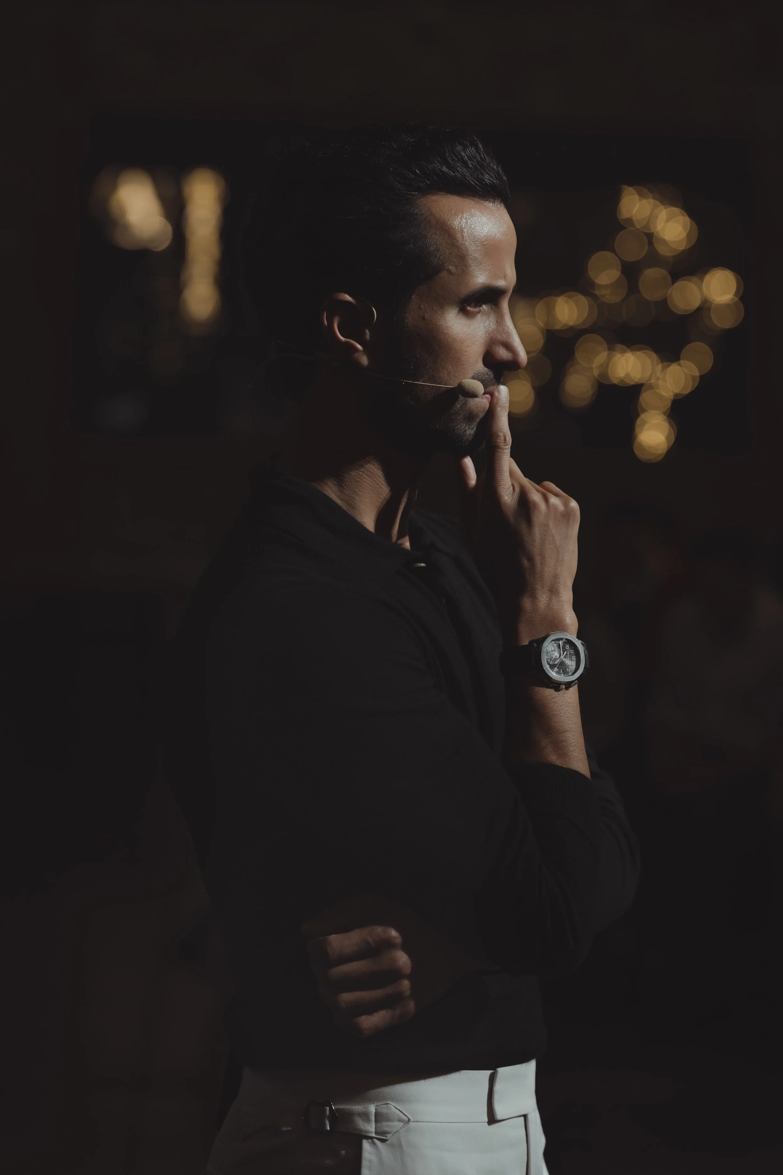 Side view of a man in a dark setting, wearing a microphone headset and a watch, with blurred warm lights in the background.