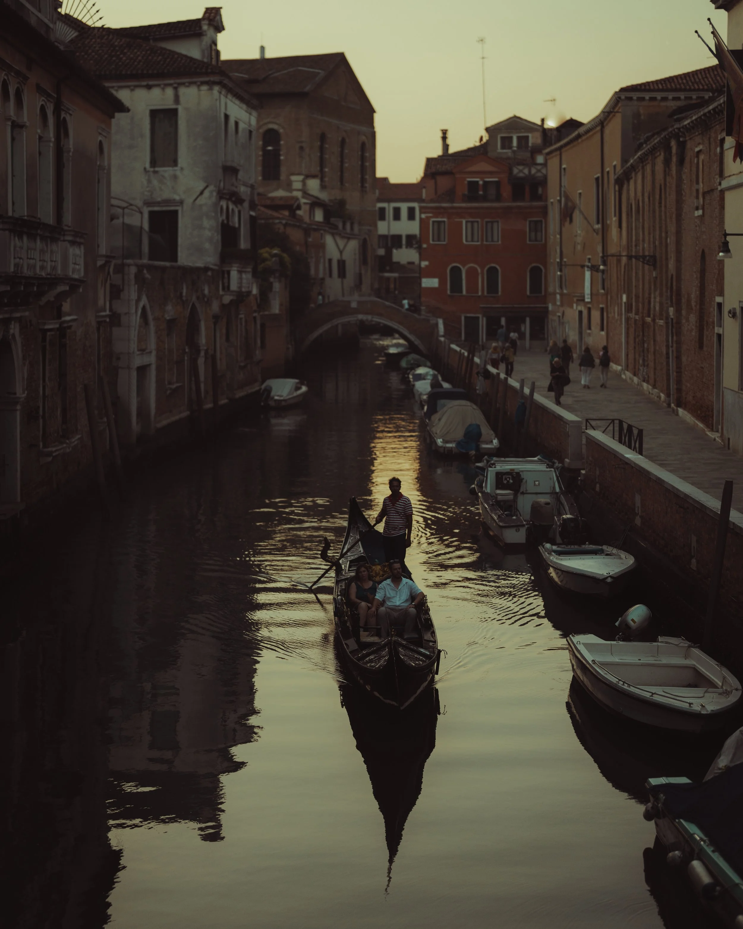 A canal scene in Venice at sunset with a gondola carrying three people and a gondolier, lined with boats docked along the sides, historic buildings, and a small pedestrian bridge in the background.