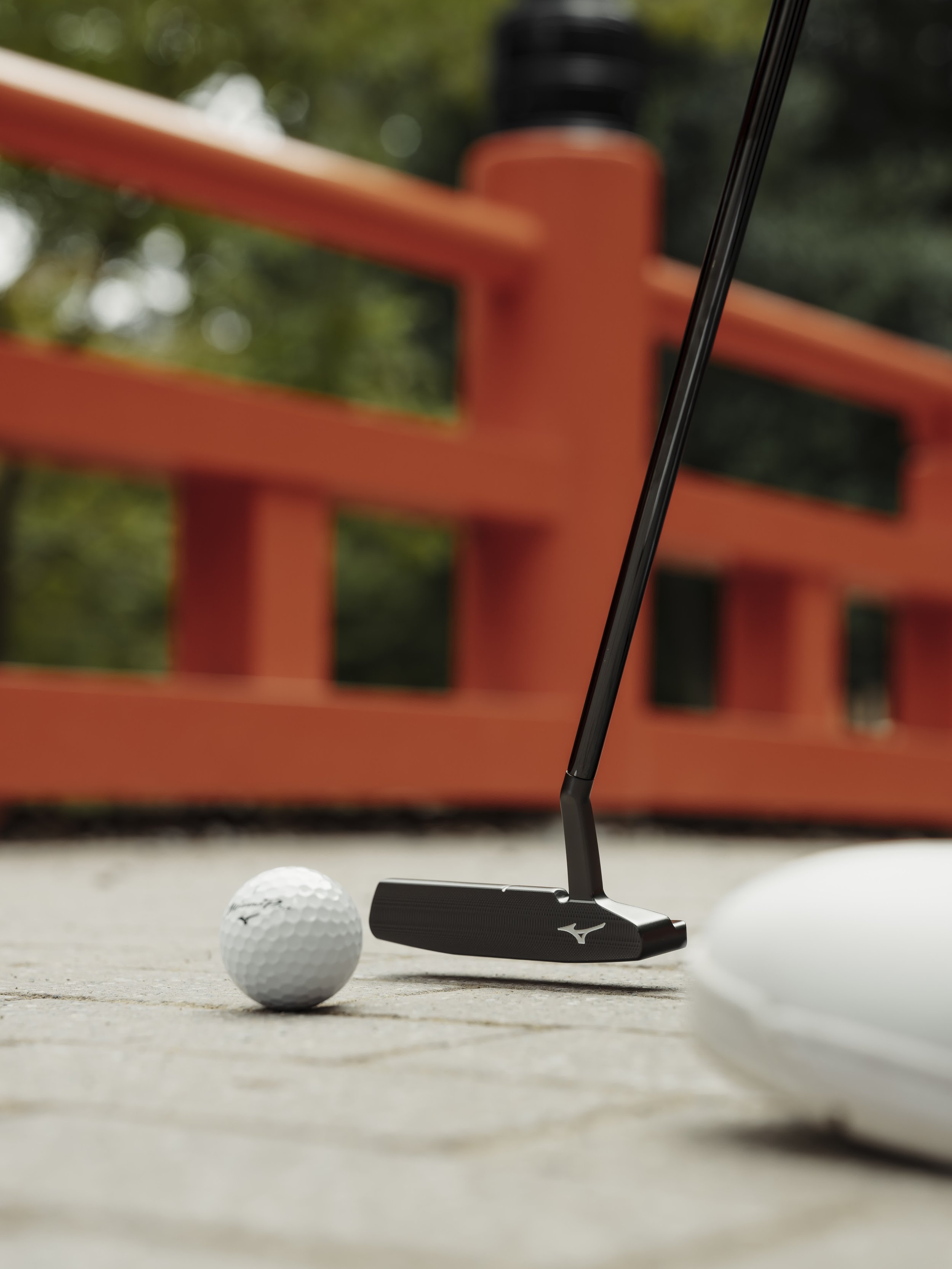 Close-up of a golf club preparing to hit a golf ball on a golf cart path, with a red wooden bench and green trees in the background.