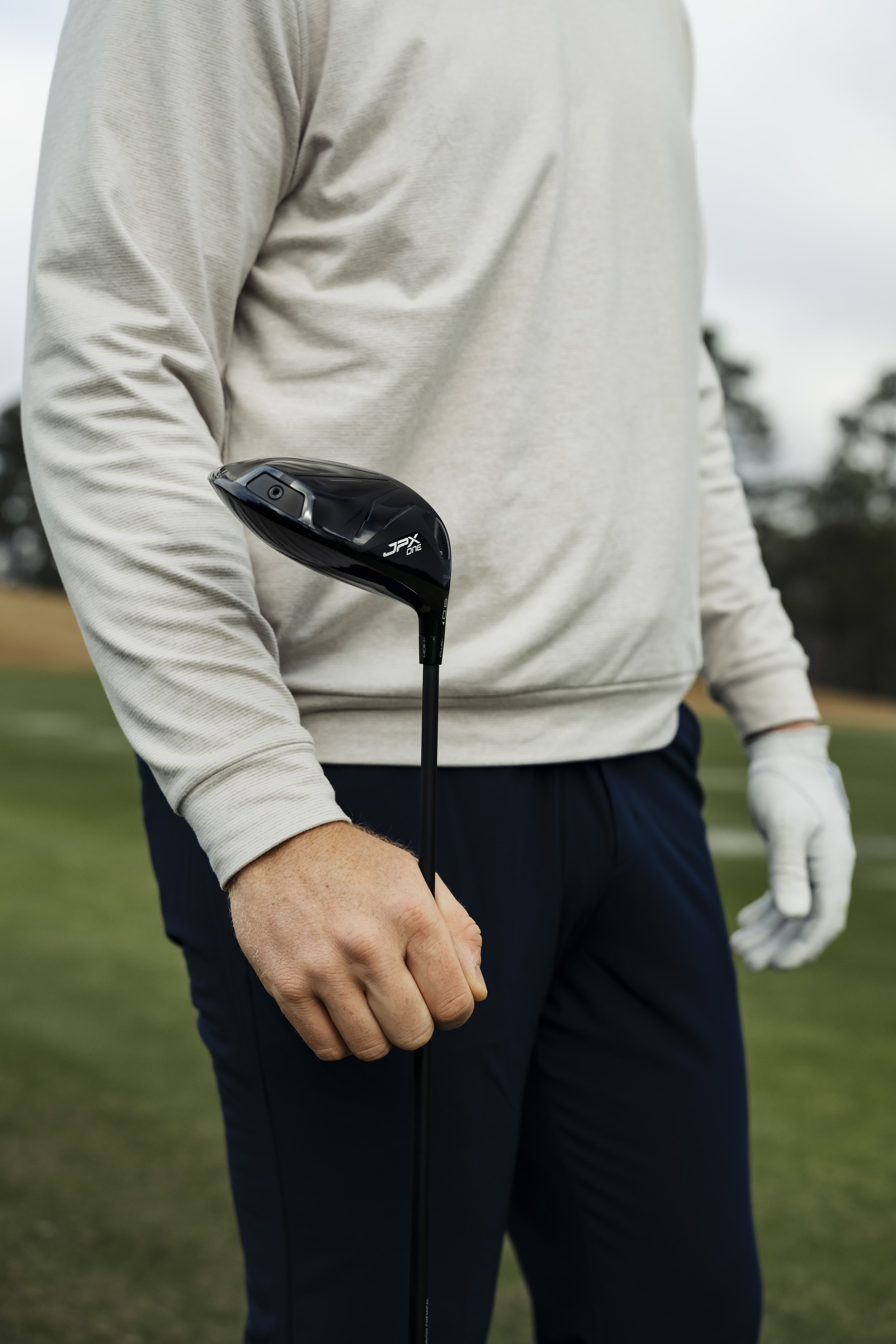 Person holding a black golf club on a golf course, wearing a light-colored long sleeve shirt and dark pants.