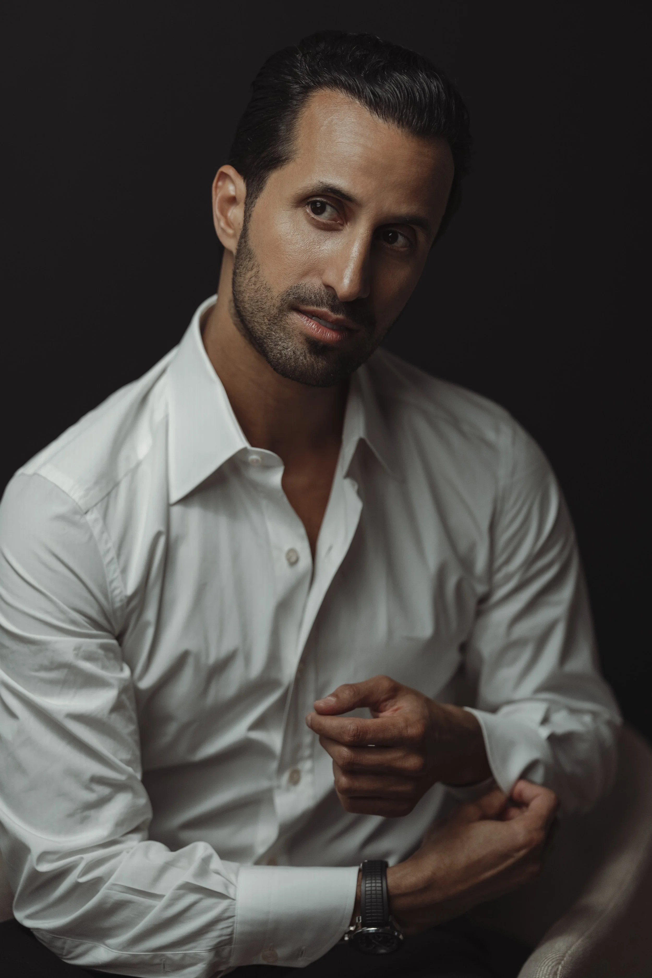 A man with dark hair, dressed in a white shirt, is adjusting his cuffs against a dark background.