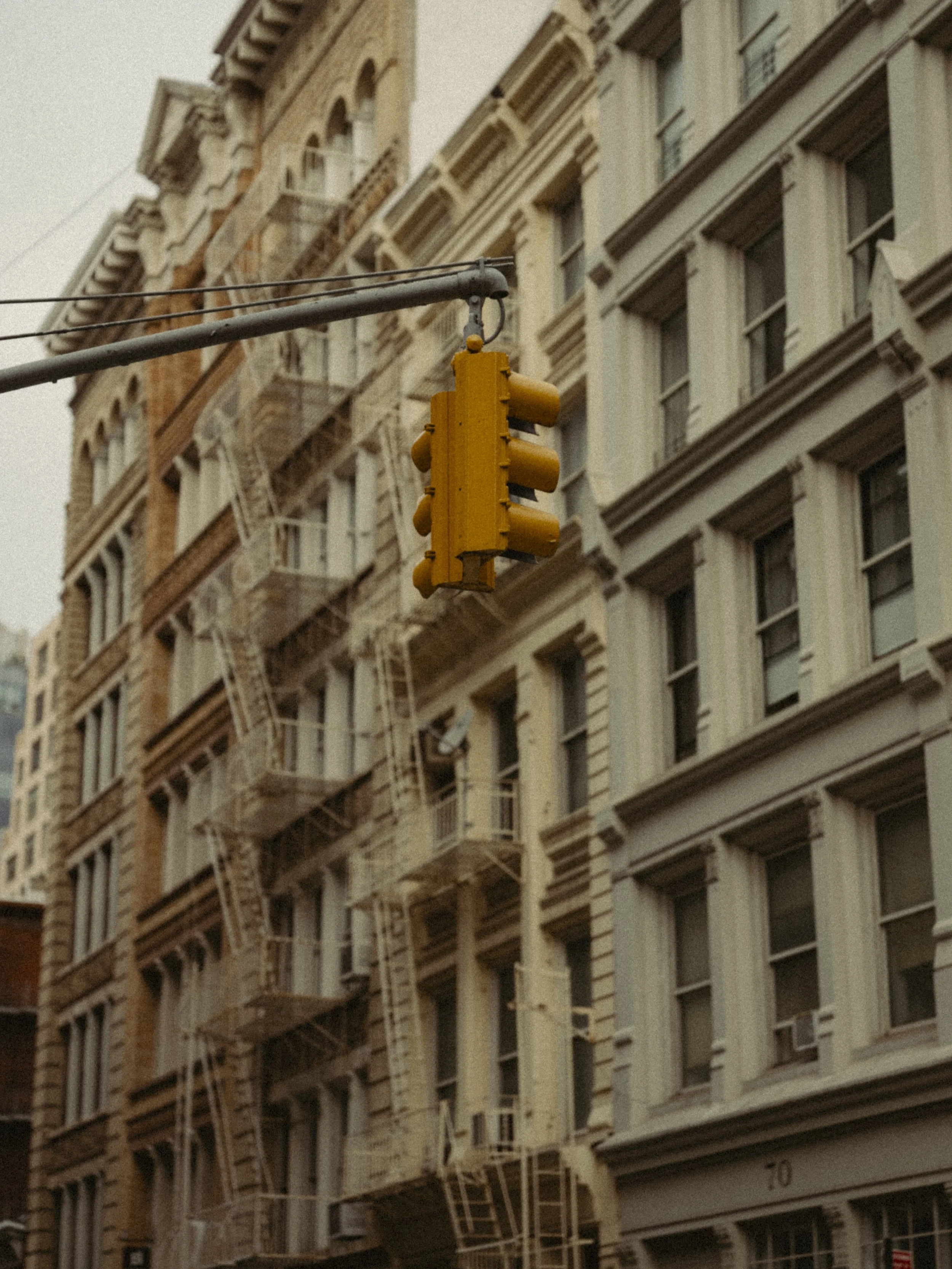 Yellow traffic light hanging over a city street with tall buildings and fire escapes.