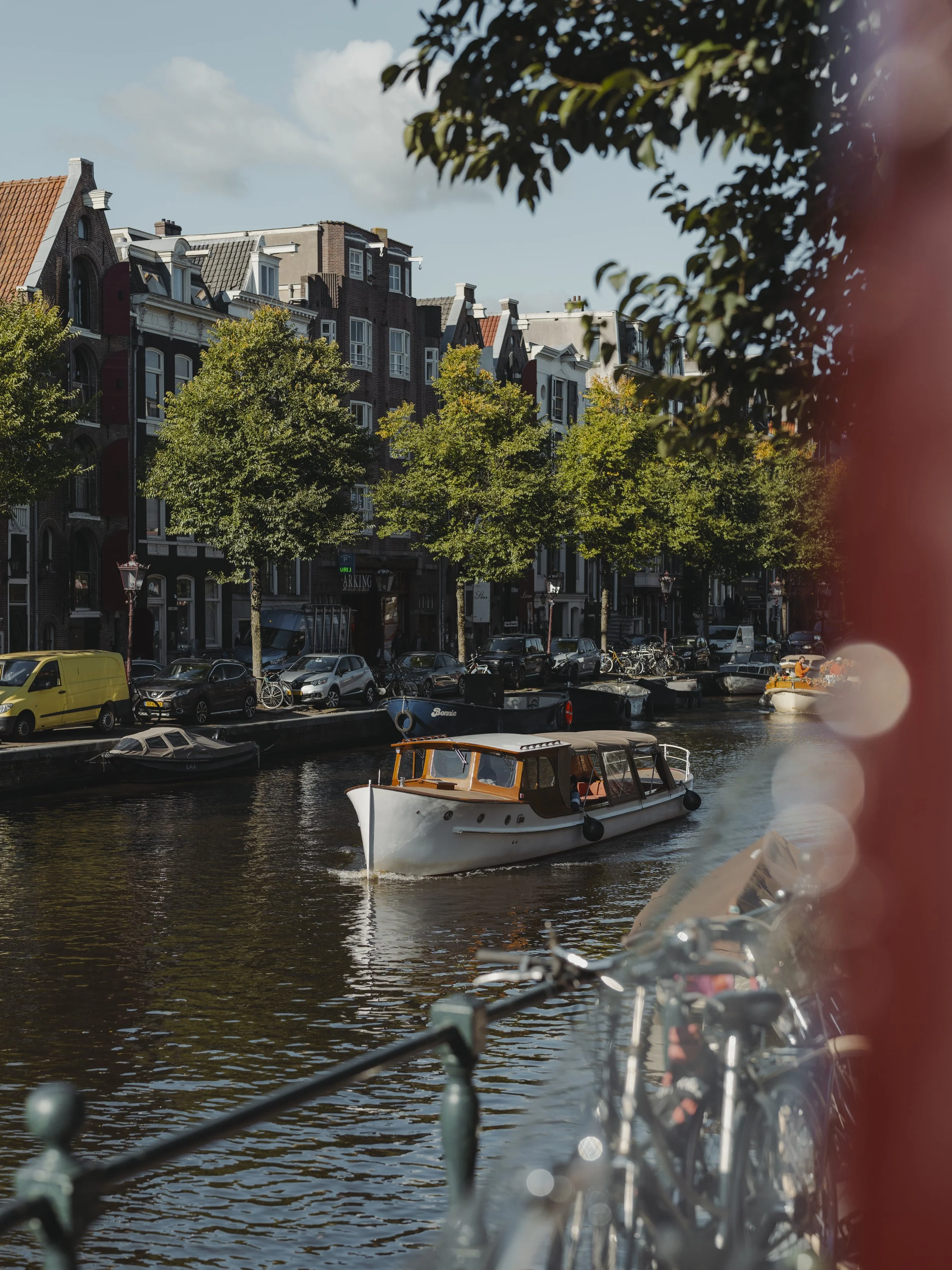 A canal scene with a white boat, parked cars, bicycles, and historic brick buildings with trees along the bank under a partly cloudy sky.