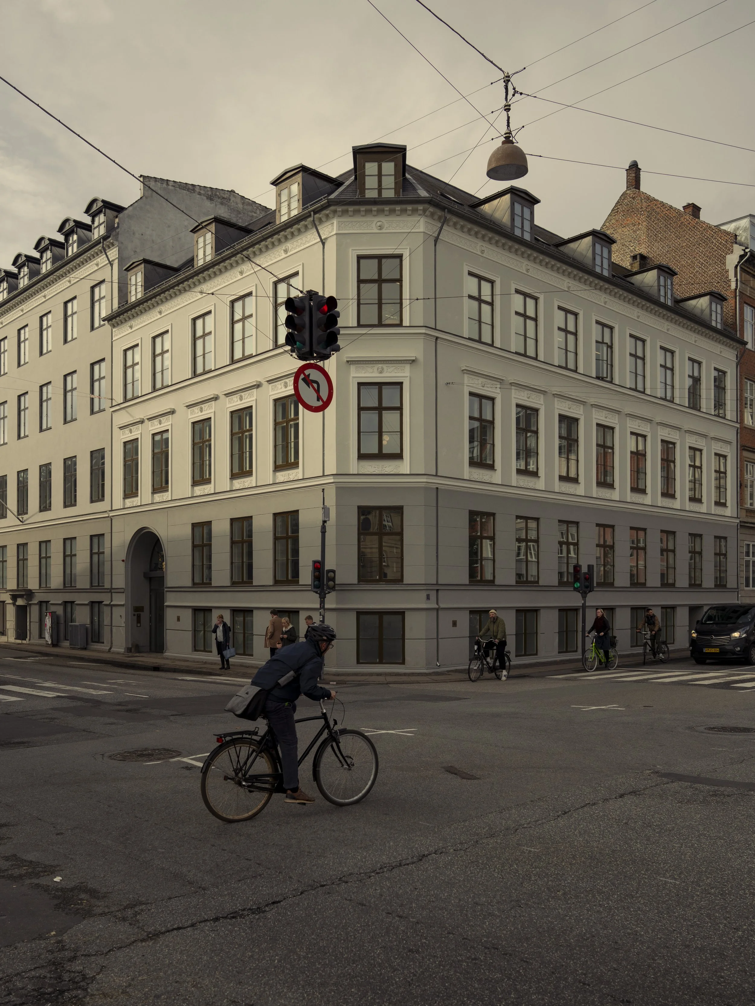City street corner with a white multi-story building, traffic lights, and pedestrians, some biking.