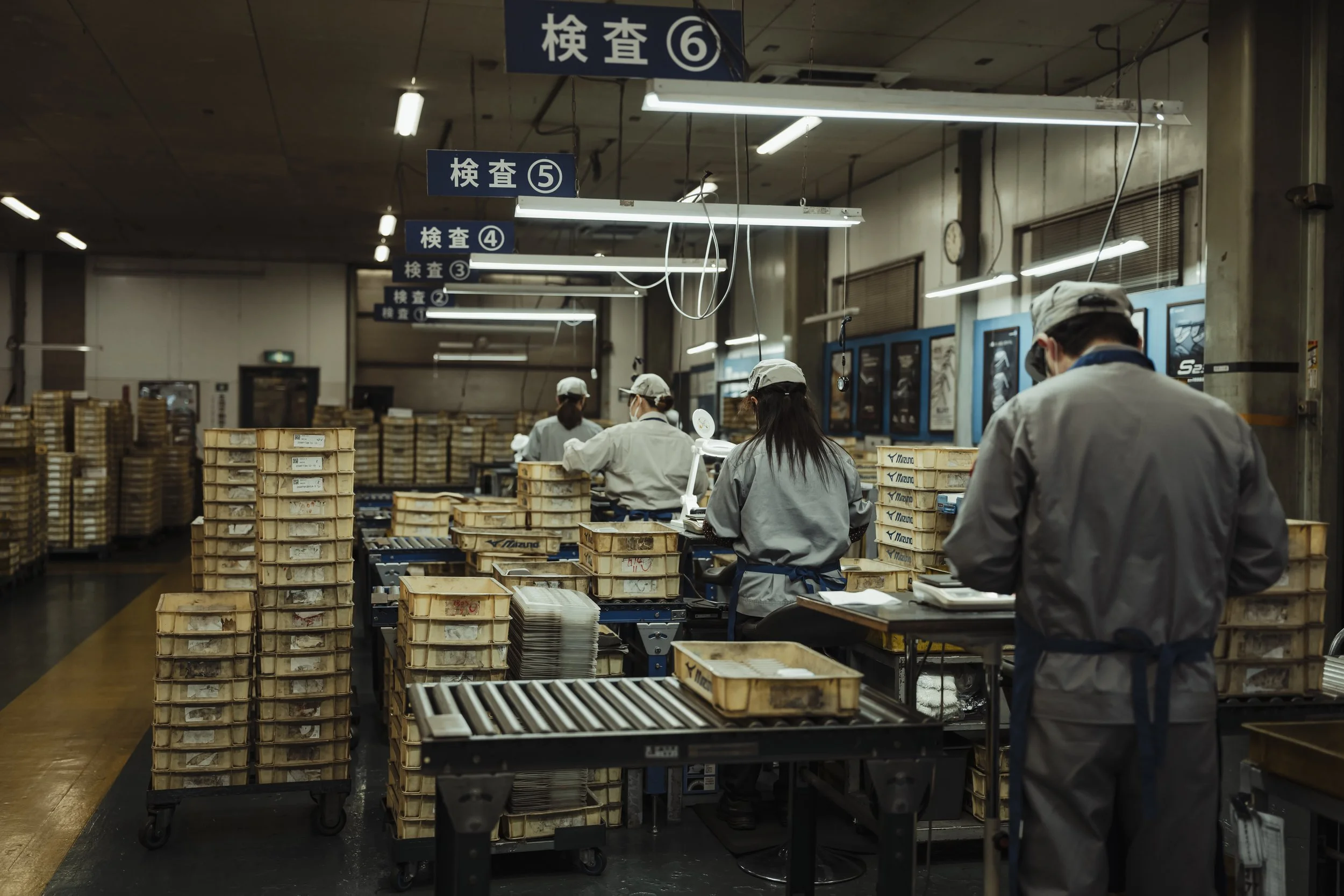 Workers in a factory assembly line with numerous stacked boxes, working under fluorescent lighting, wearing uniforms and caps, with signs overhead in Japanese and operating equipment.