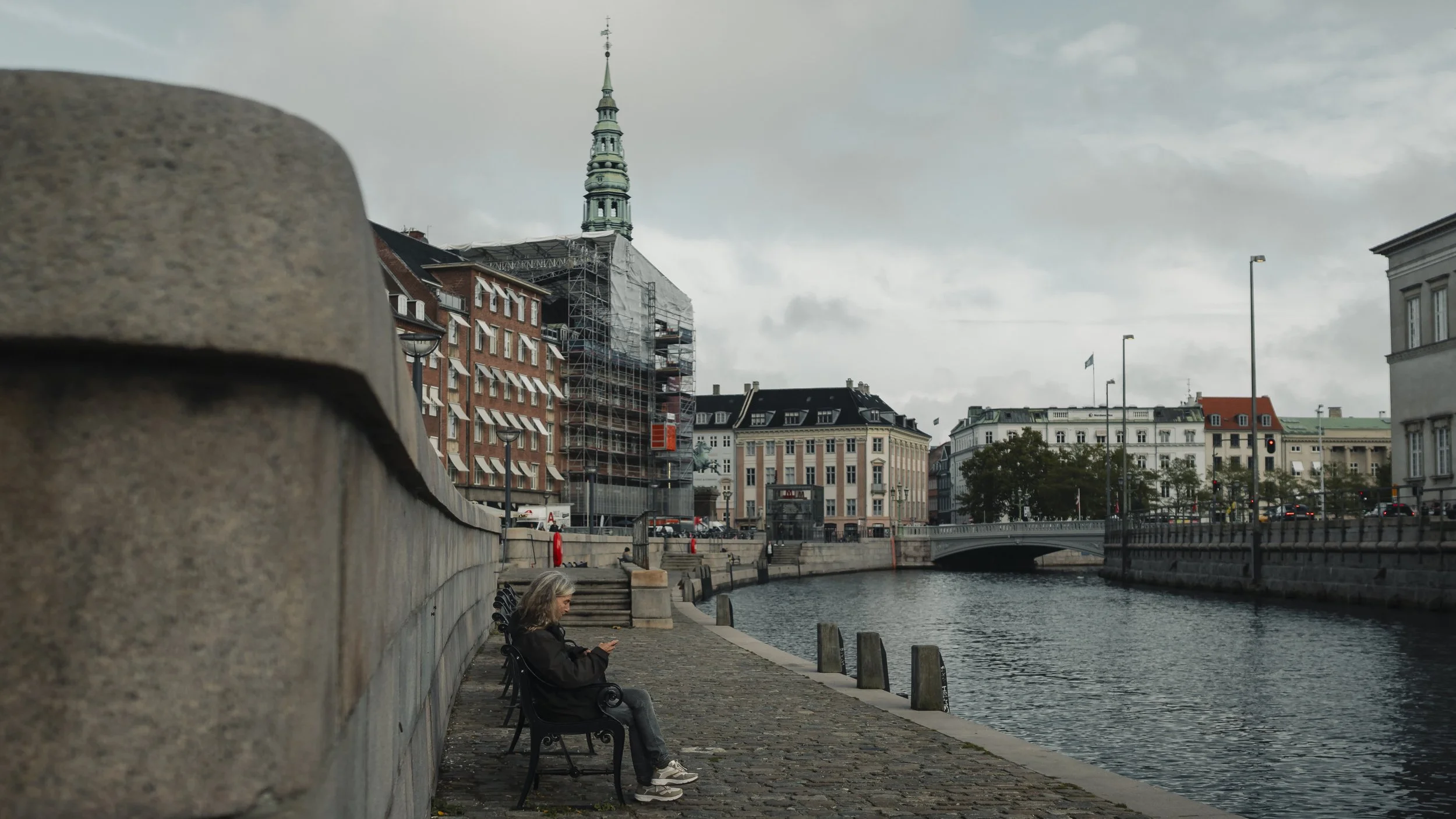 A woman sitting on a black park bench by a river, looking at her phone, with city buildings and a bridge in the background under a cloudy sky.
