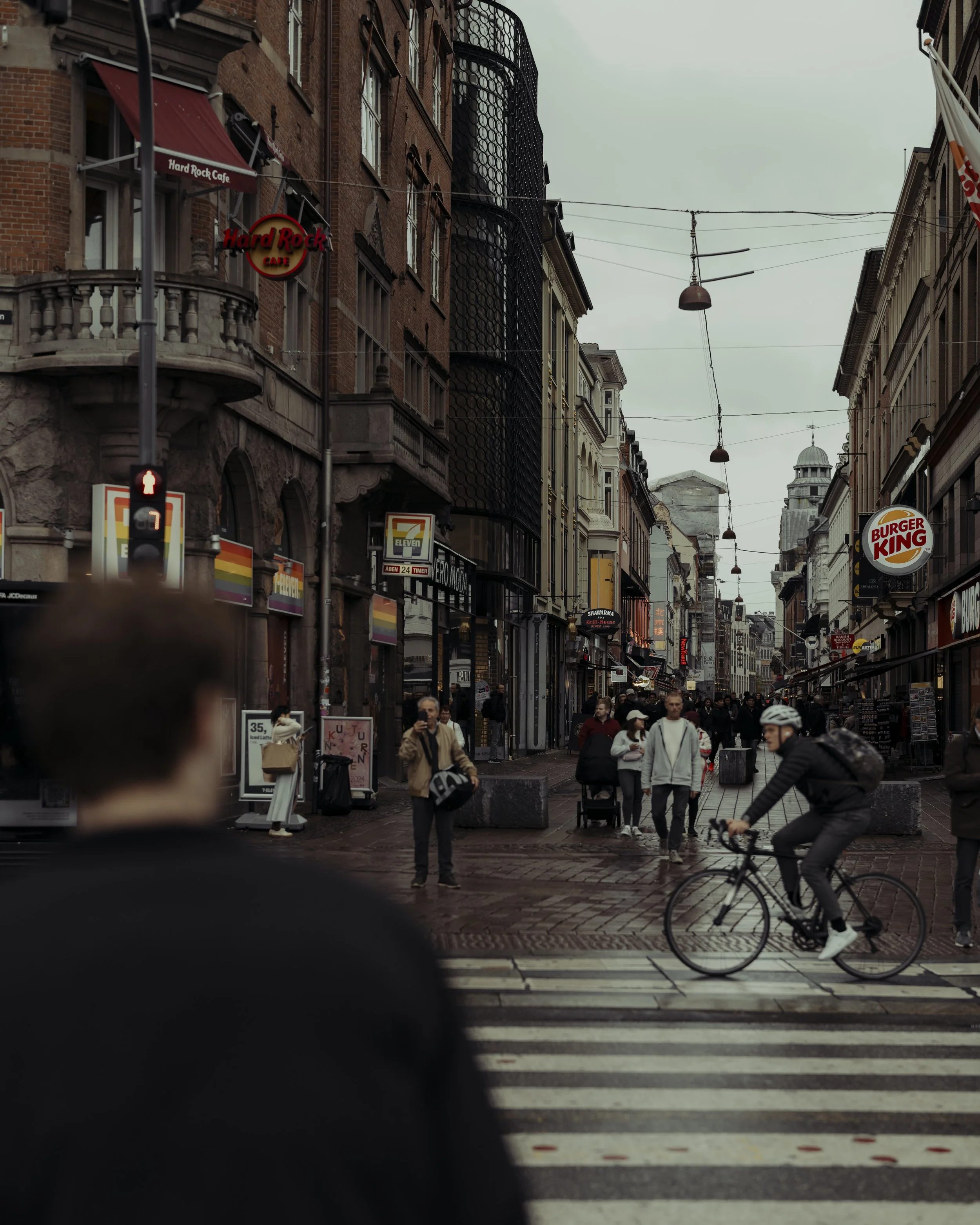 A city street scene with pedestrians and a cyclist crossing a crosswalk. There are various storefronts, signs including Hard Rock Cafe and Burger King, and buildings lining the street under an overcast sky.