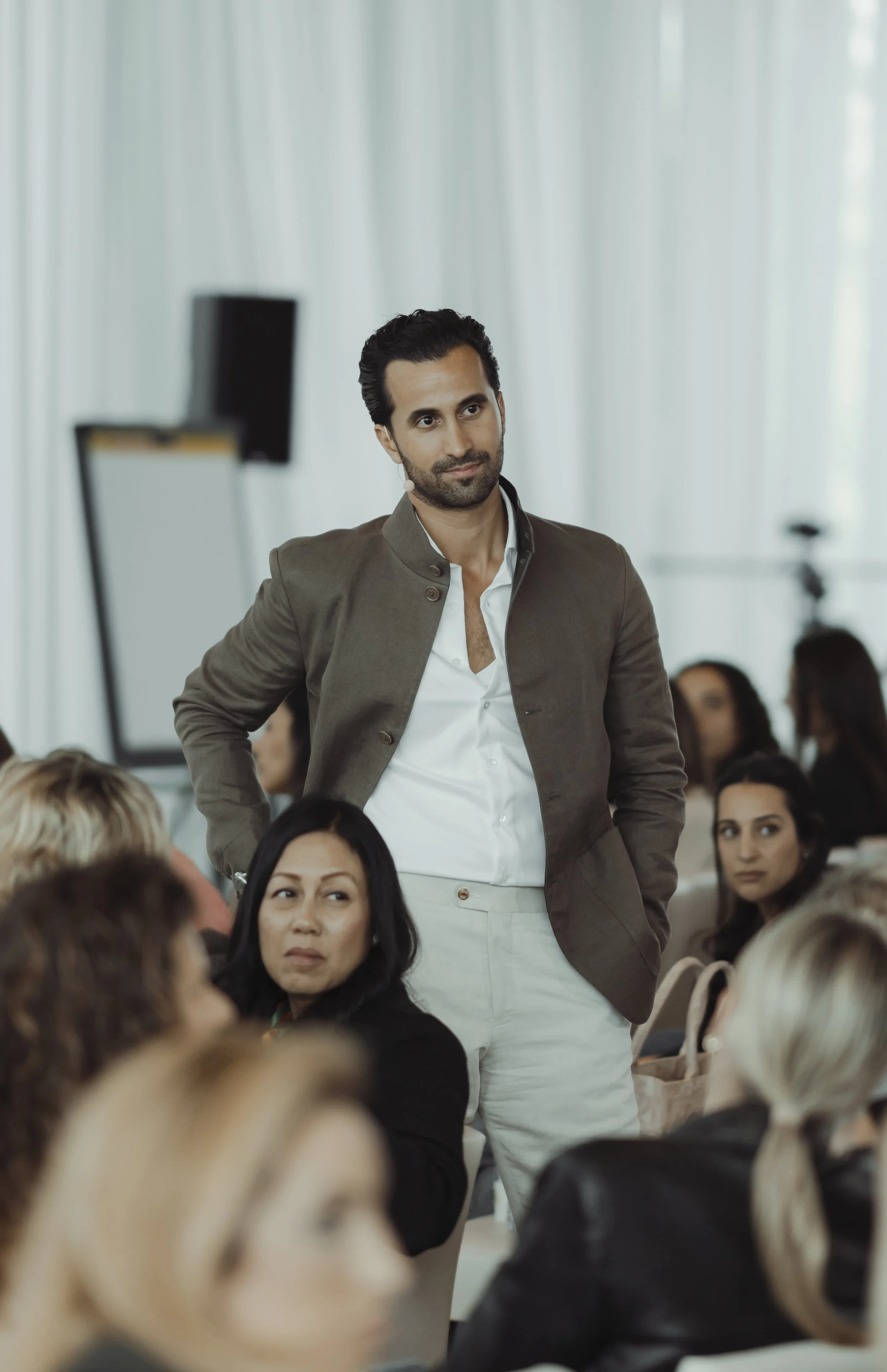 A man standing with his hands on his hips during a conference or seminar, surrounded by seated attendees in a bright room.