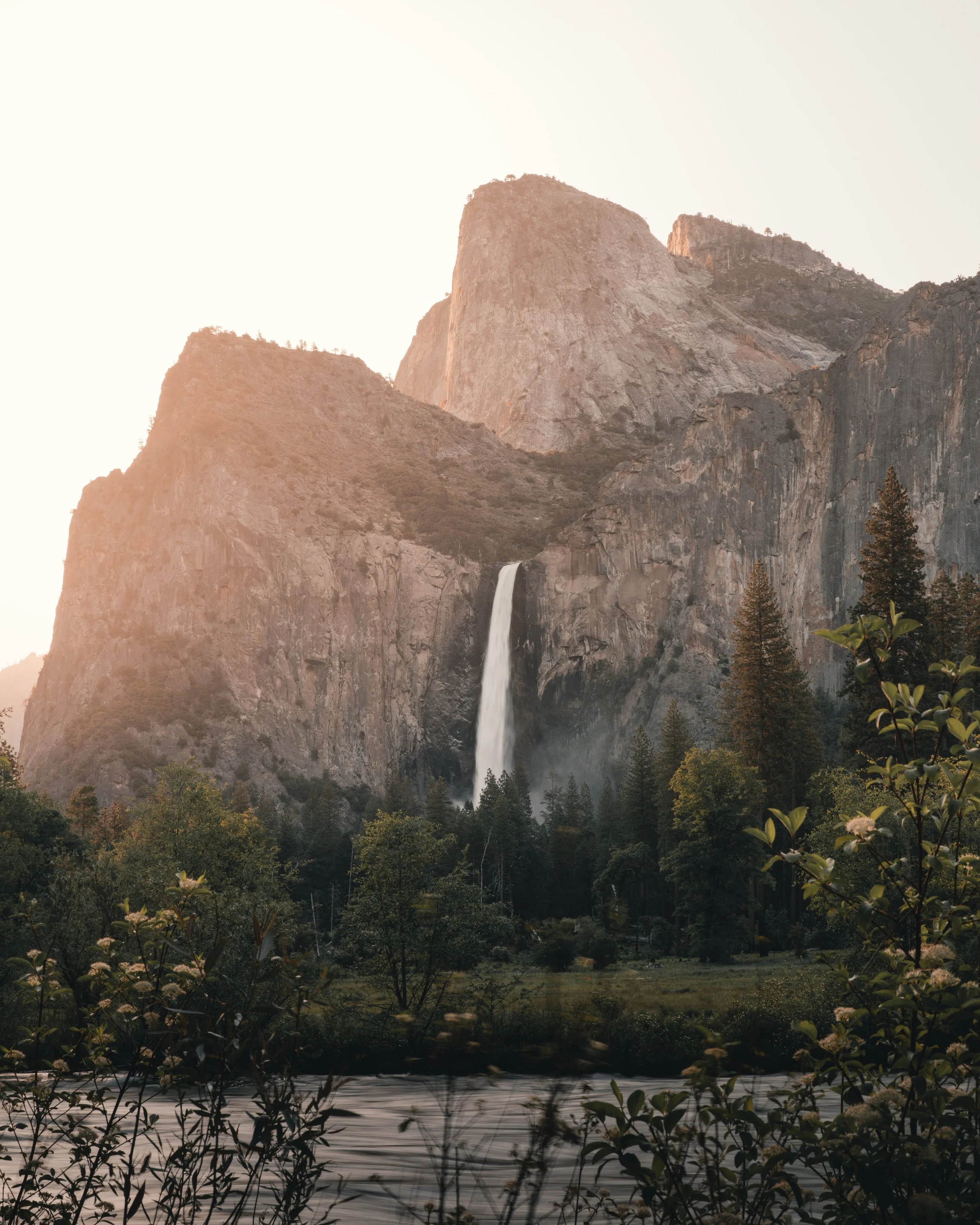 Scenic landscape with large rocky mountain, waterfall, dense forest, and river at sunset.