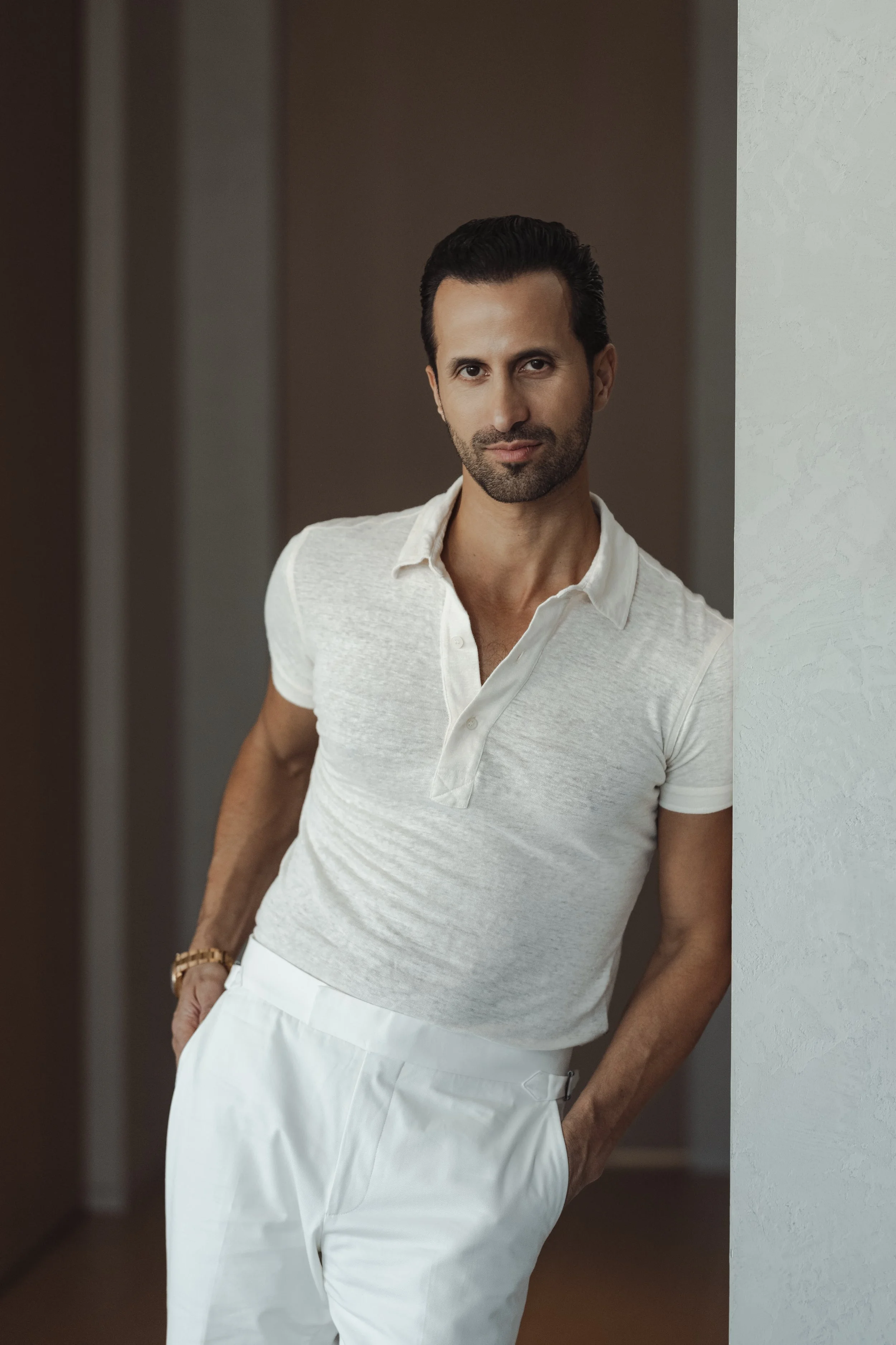 A man leaning against a wall, wearing a light-colored polo shirt and white pants, with a neutral background.