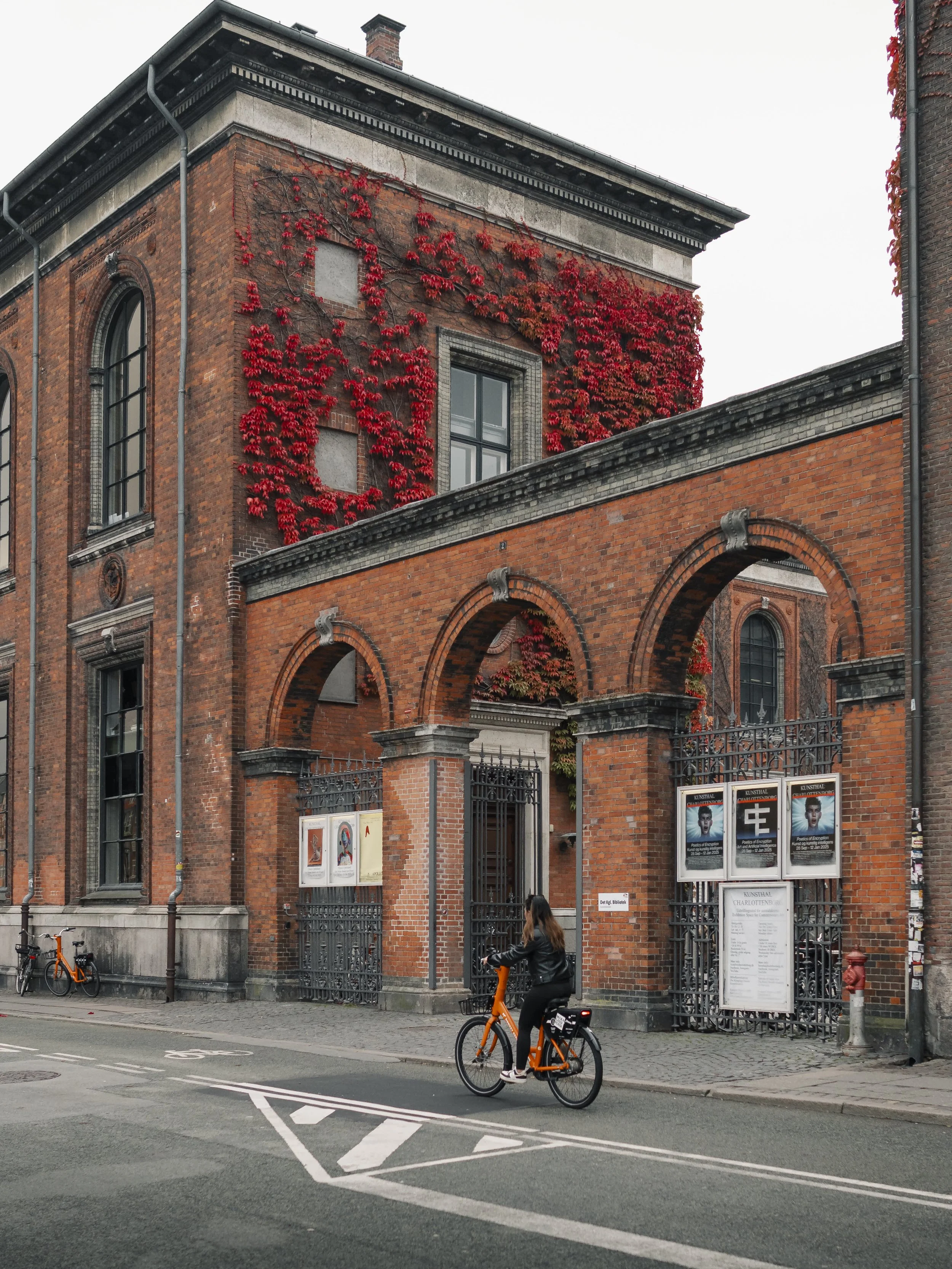 A woman riding a bright orange bicycle past a historic red brick building with arched doorways and windows, some of which are covered with red vines, on a city street.