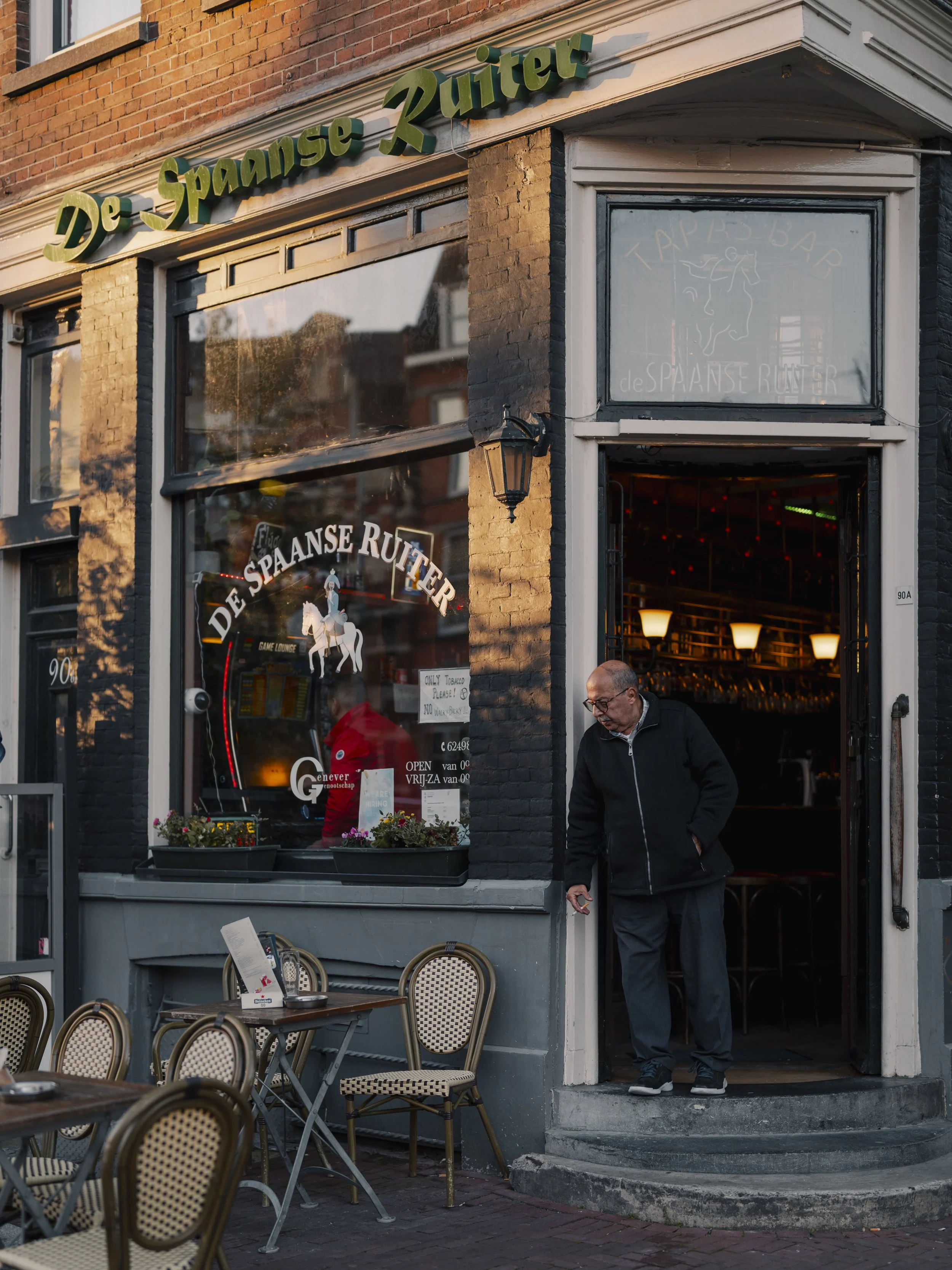 A man in black jacket and glasses walking out of a Spanish restaurant called De Spaanse Ruiter, with outdoor tables and chairs in front.