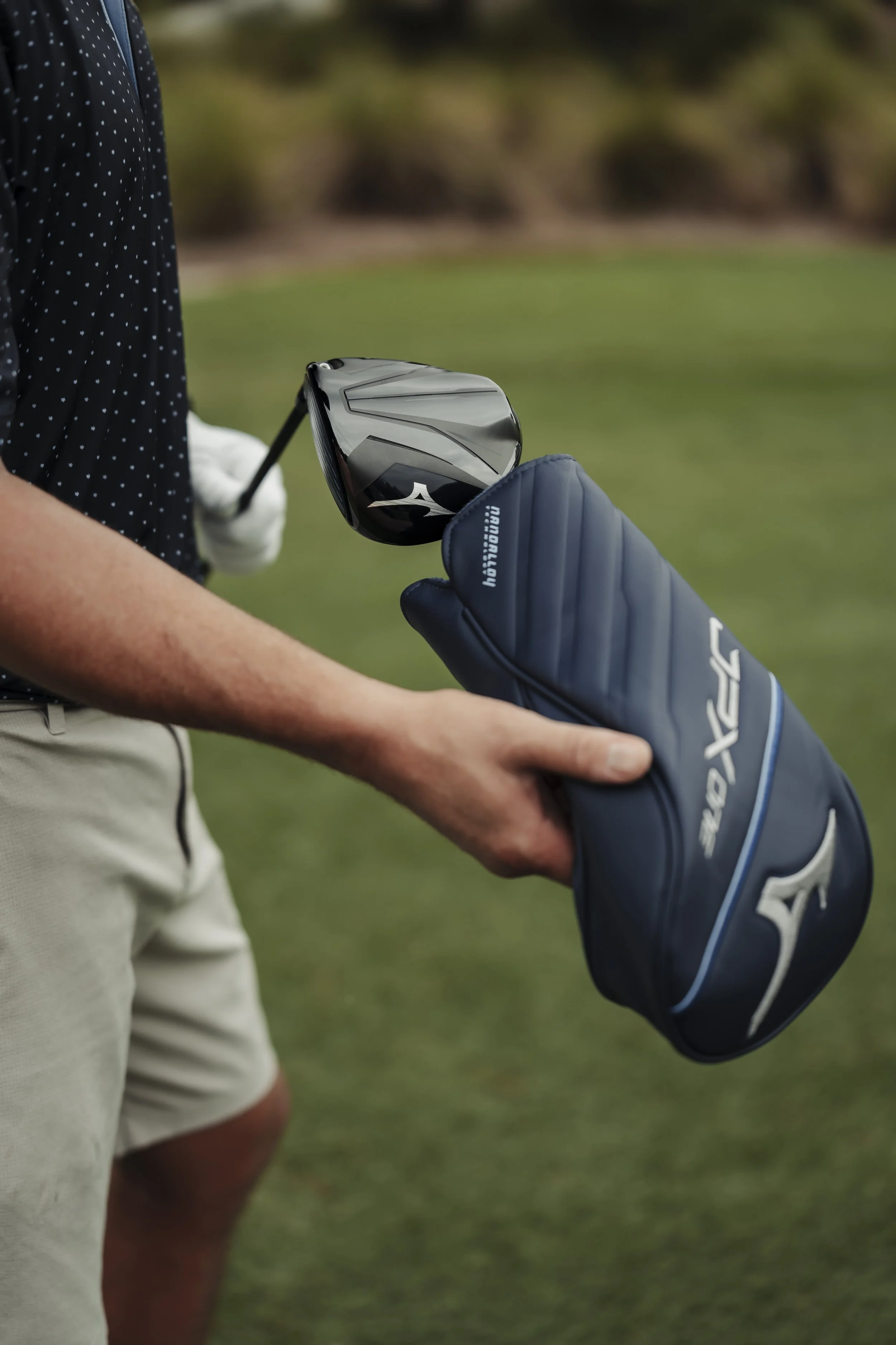 A person holding a golf club and a blue Mizuno golf headcover on a golf course.
