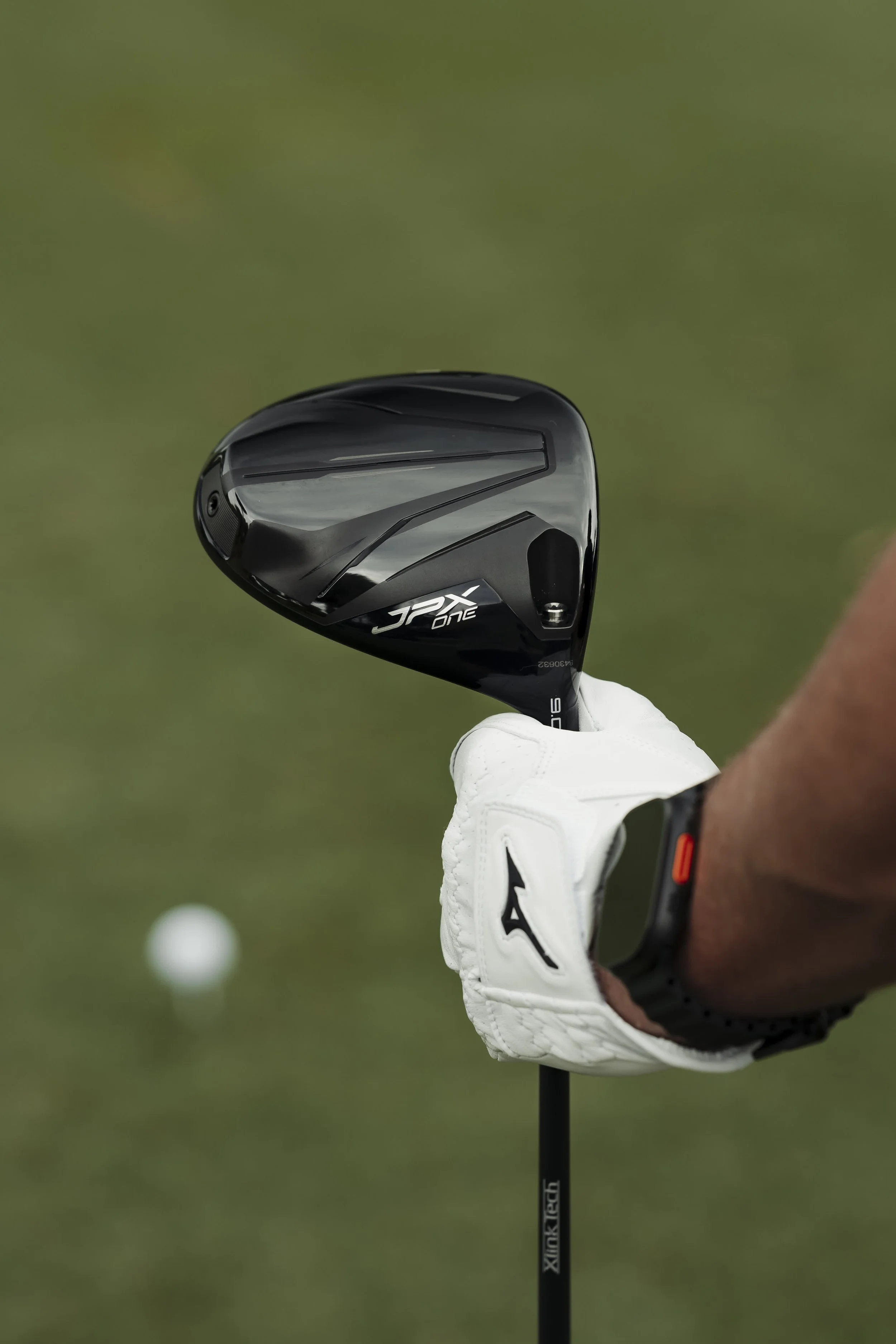 Close-up of a golf club, held by a golfer wearing a white glove on ungloved hand, with a golf ball in the background on a grassy course.