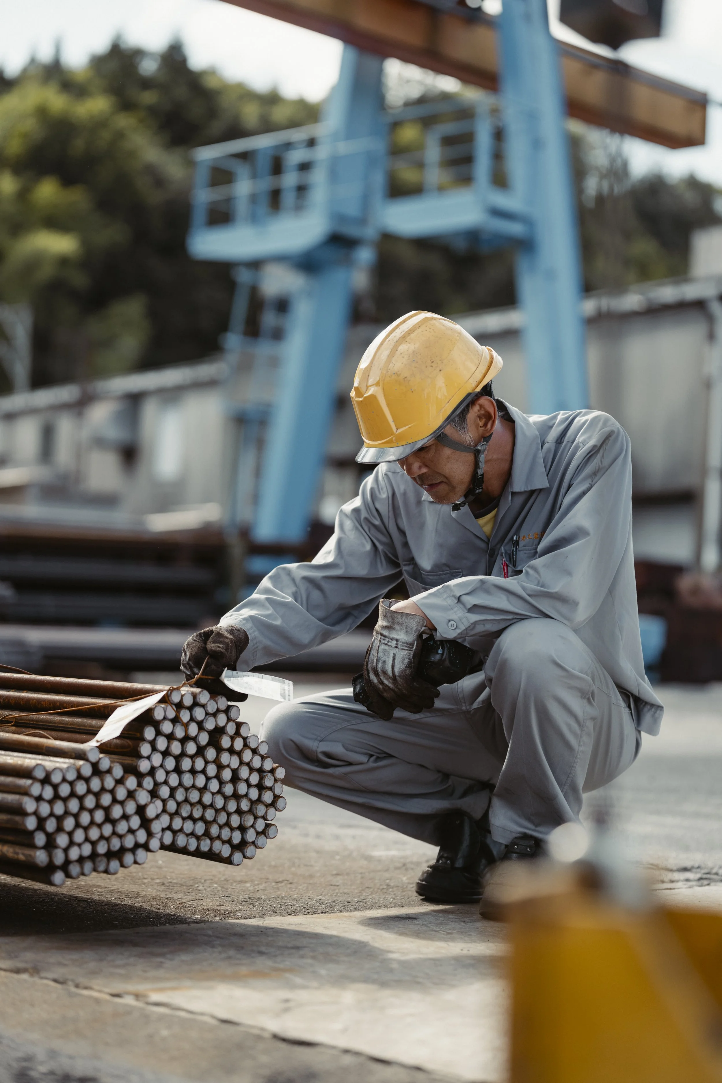 A male construction worker wearing a yellow safety helmet and gray work clothes, inspecting metal rods at a construction site.