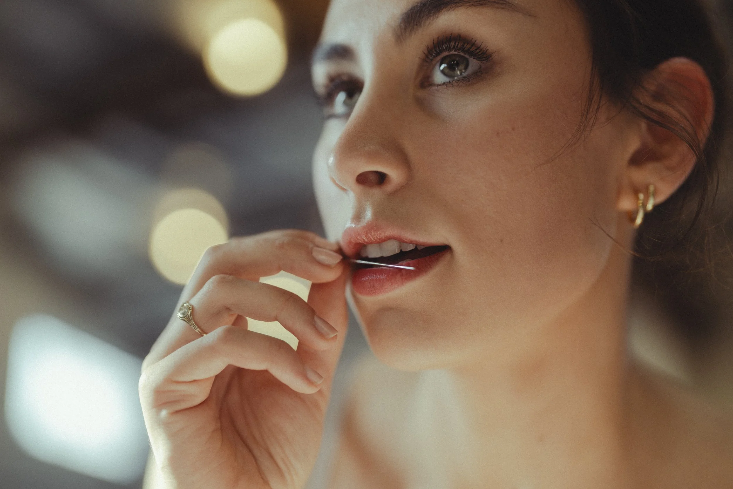 A close-up of a young woman applying lipstick, with blurred lights in the background, wearing gold earrings and a ring.