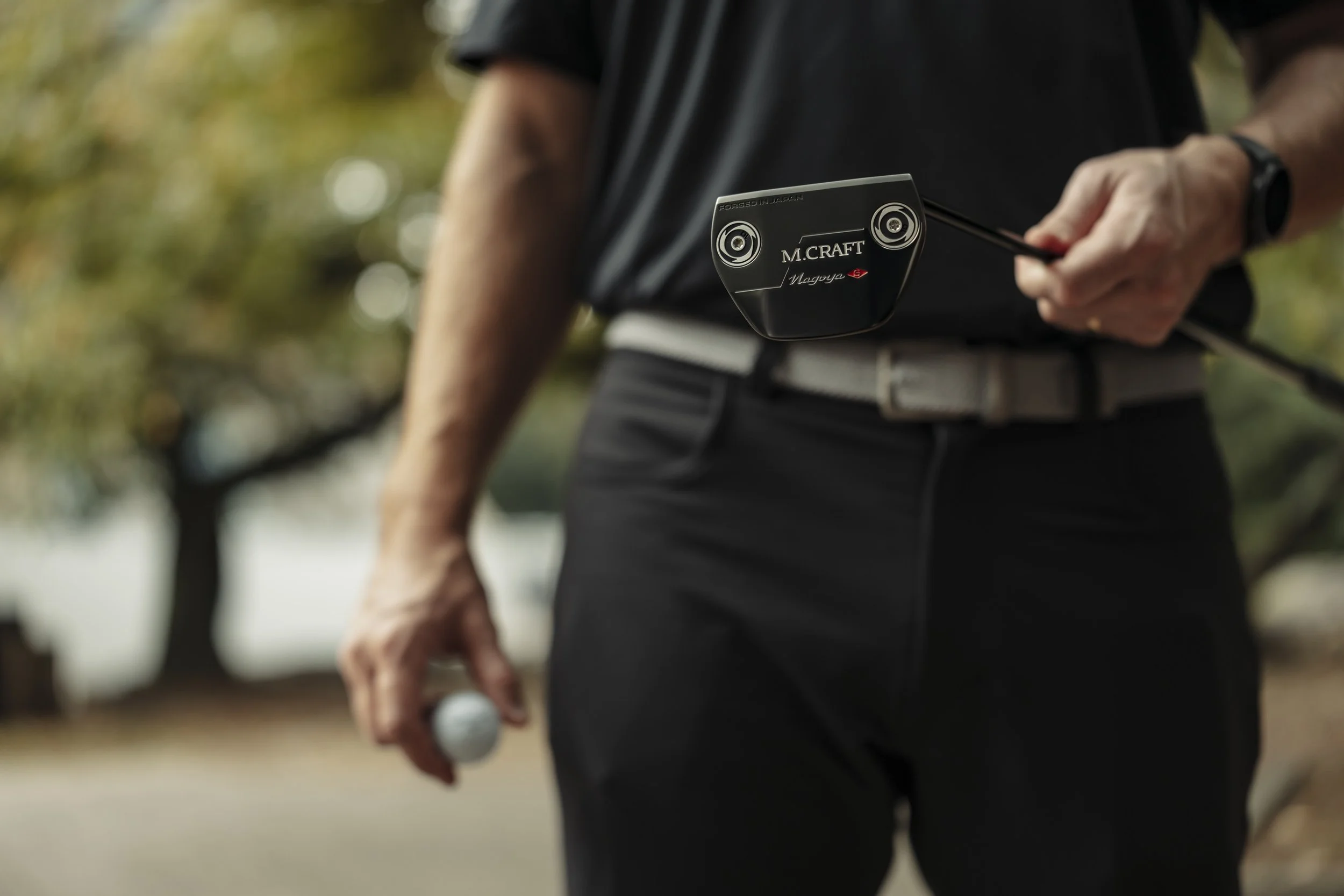 A person dressed in black holding a golf club and a golf ball outdoors, with trees in the background.
