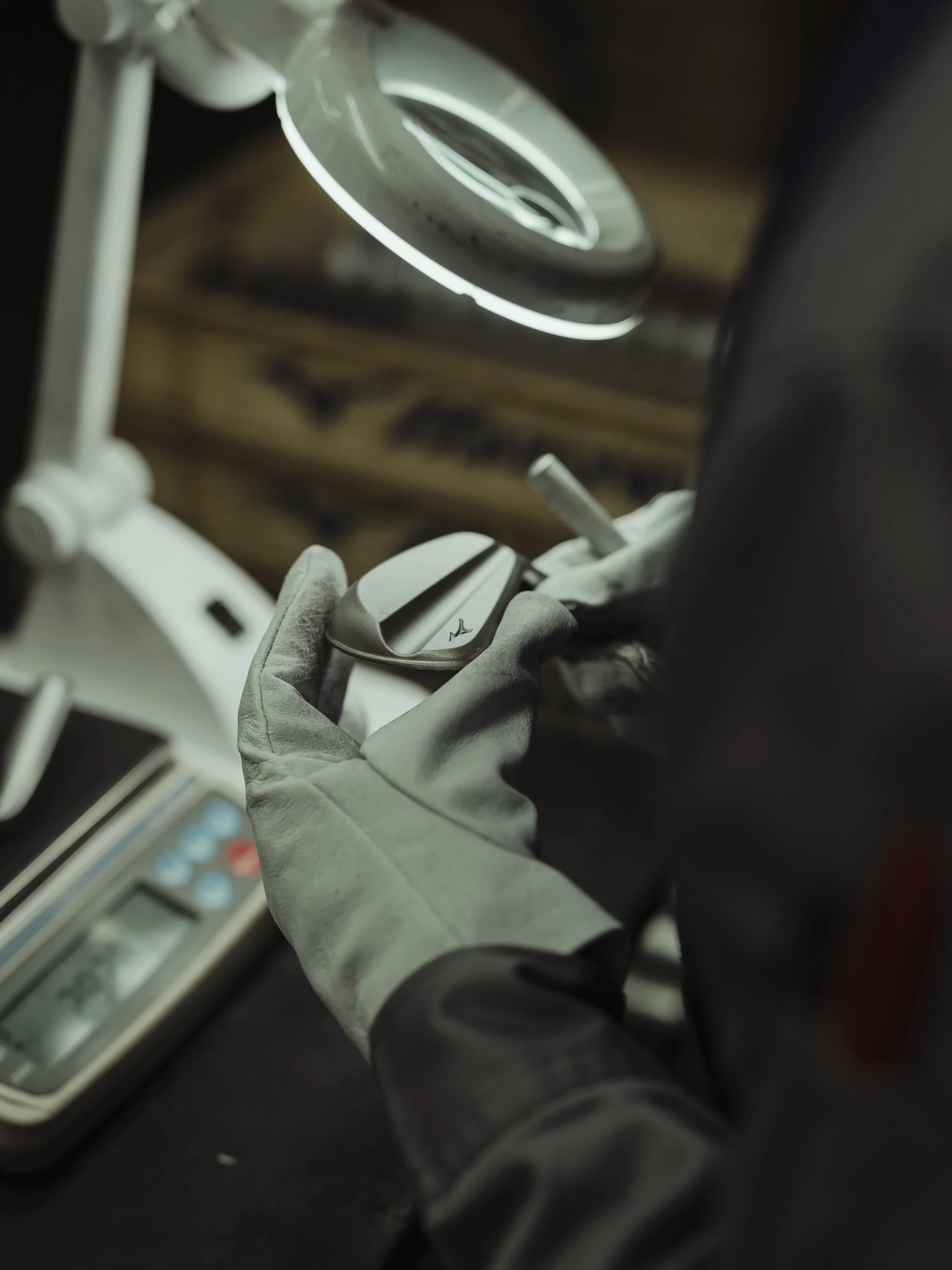 Close-up of a worker's hand wearing a gray glove, holding a computer mouse while working at a desk with a calculator in the background, under a magnifying lamp in a workshop or office setting.