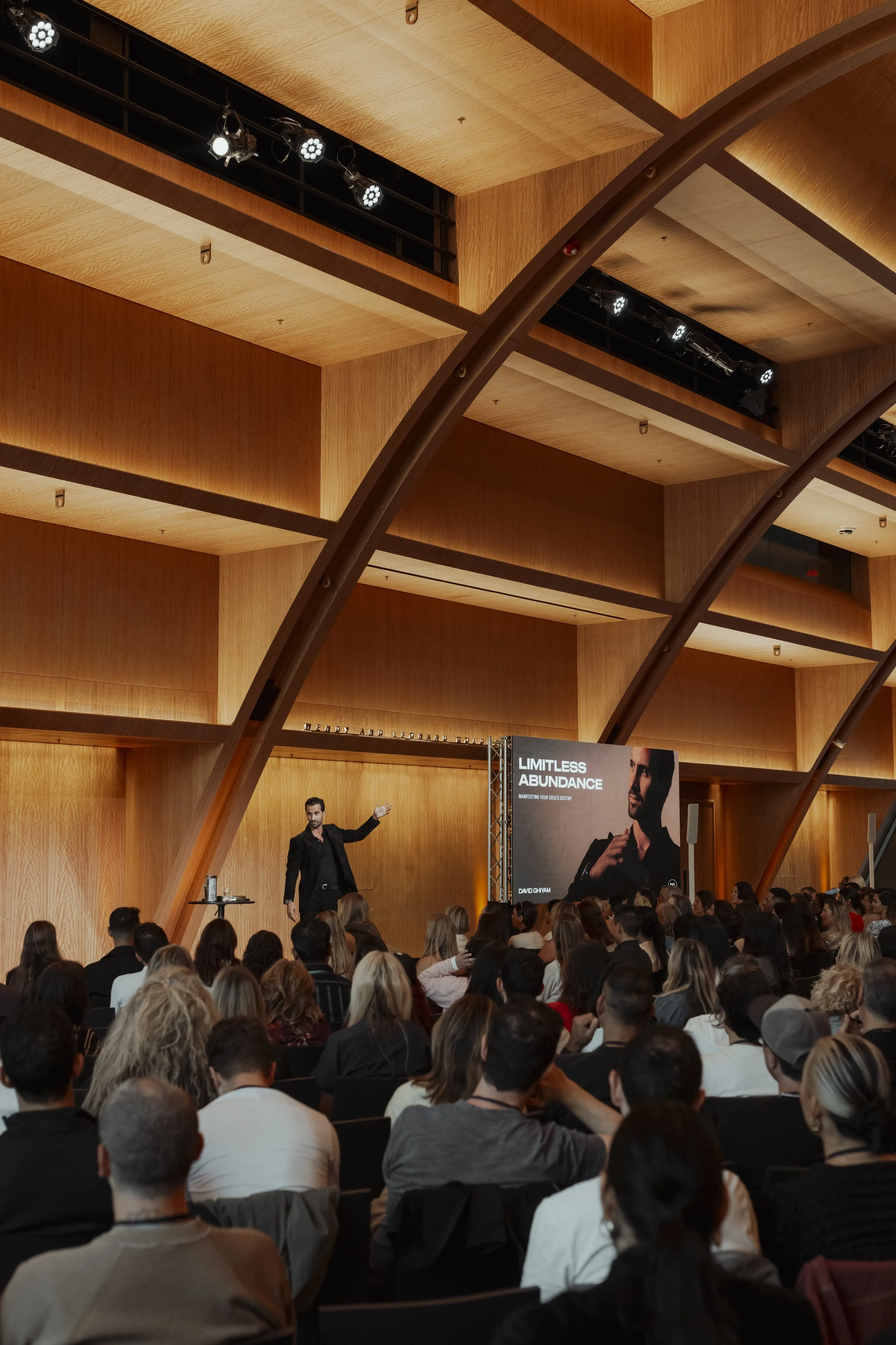 A speaker on stage at a conference, gesturing while addressing an audience, with a large screen displaying 'Limitless Abundance' and an image of a man in a black outfit.