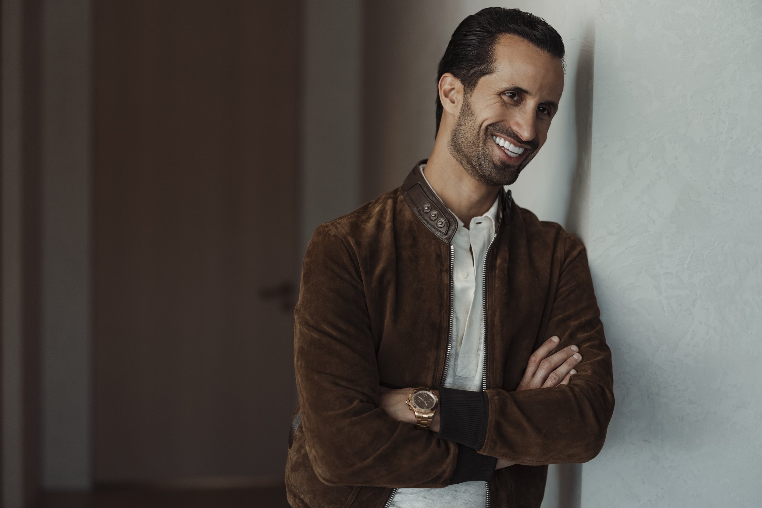 A smiling man with dark hair and beard, wearing a brown jacket and a white shirt, standing with arms crossed against a light-colored wall.