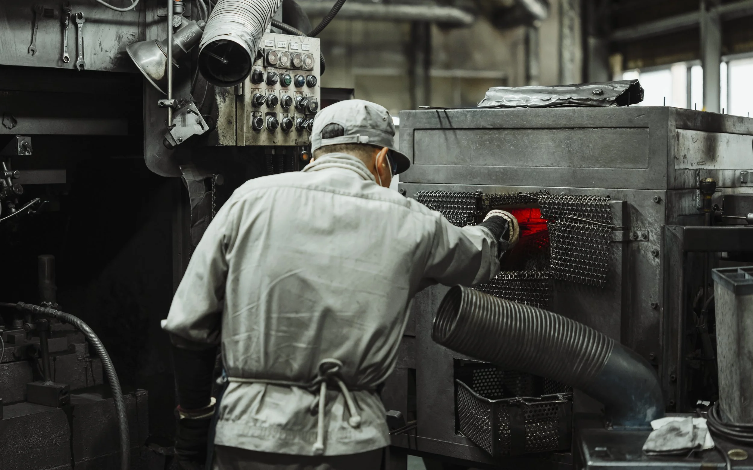 A worker wearing a gray uniform, cap, and face mask is reaching into a machine with a red glow inside.