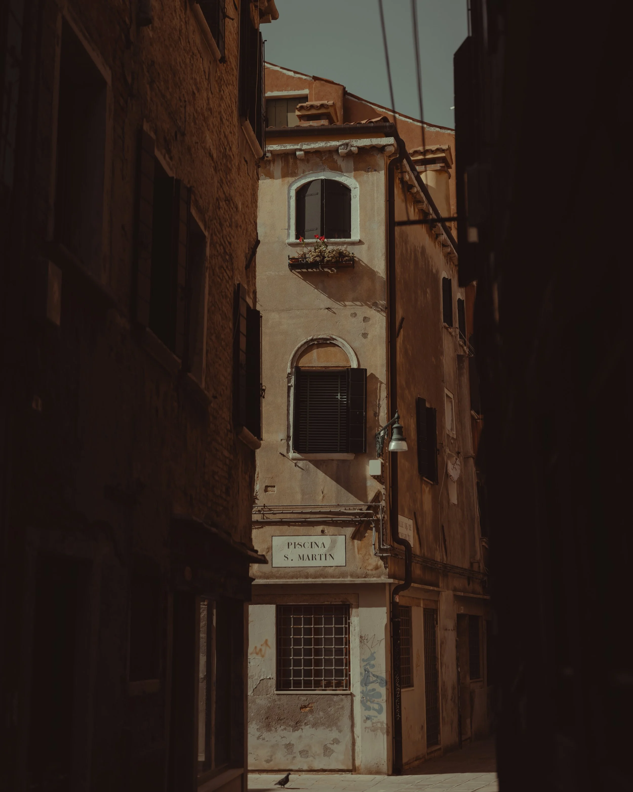 Narrow street in Italy with old, weathered buildings, open and closed shutters, a sign reading 'Piscina S. Martin', and a pigeon on the pavement.