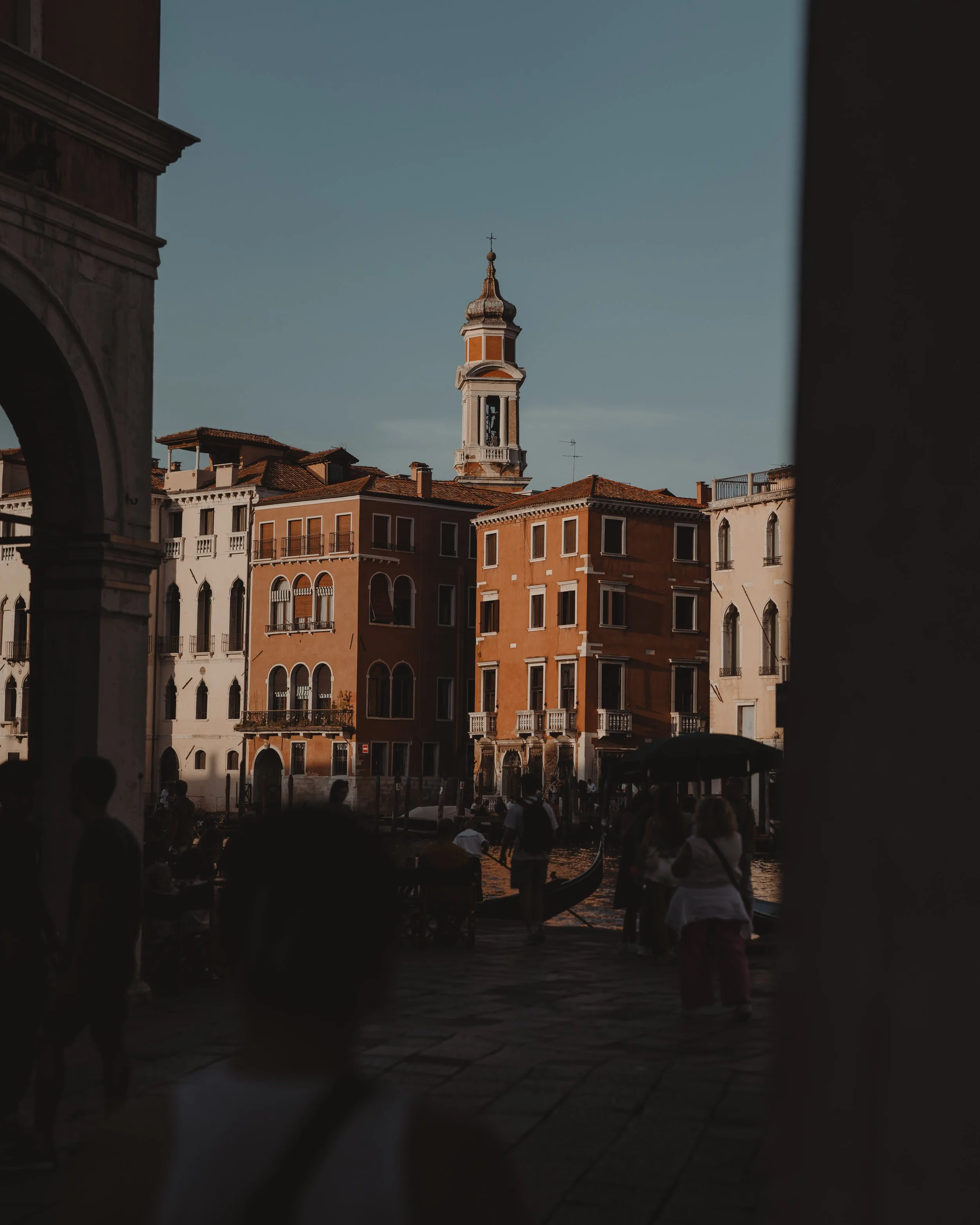 View of Venetian buildings with a bell tower and gondolas on a canal, seen through an archway, during sunset.