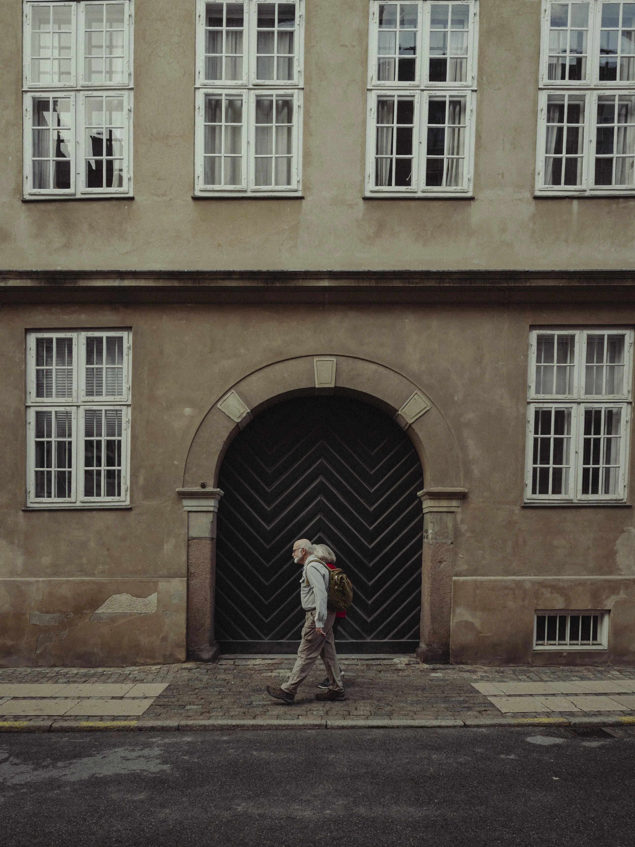 Older man with gray hair and a backpack walking past a large black doorway on a beige building with multiple windows.