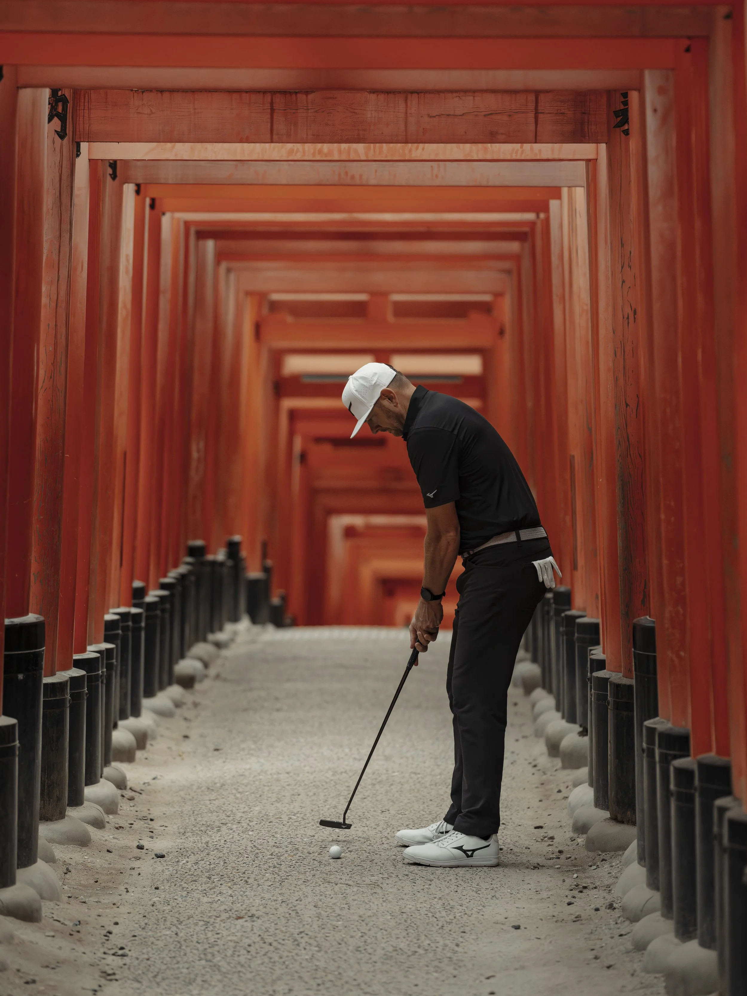 A man in black golf attire and a white cap playing golf on a path lined with red torii gates.