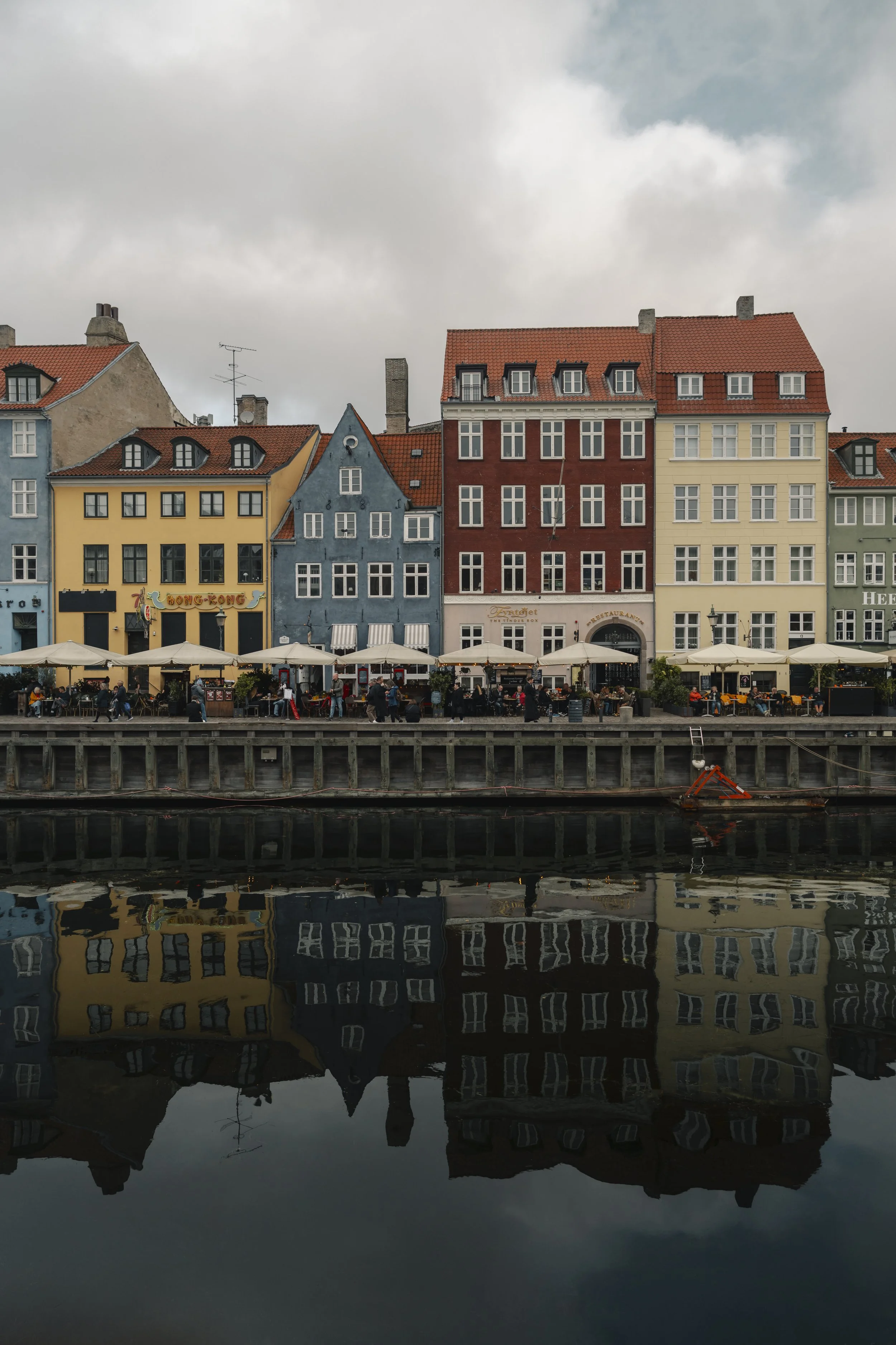 Colorful row of buildings along a waterfront with outdoor cafes and people dining, with their reflections visible in the water.