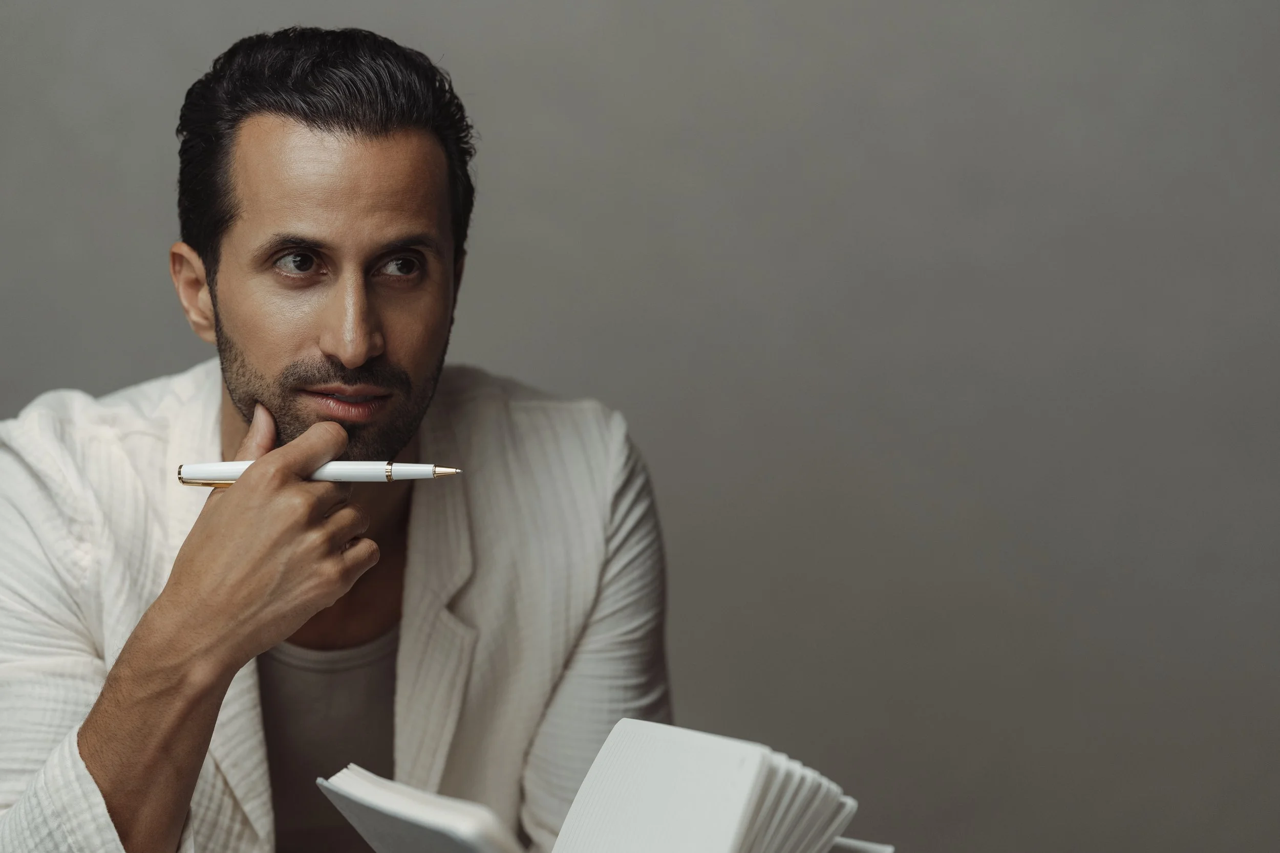 A man with dark, short hair and a beard, wearing a white shirt, is holding a white pen near his mouth while looking thoughtfully to the side, with a gray background.