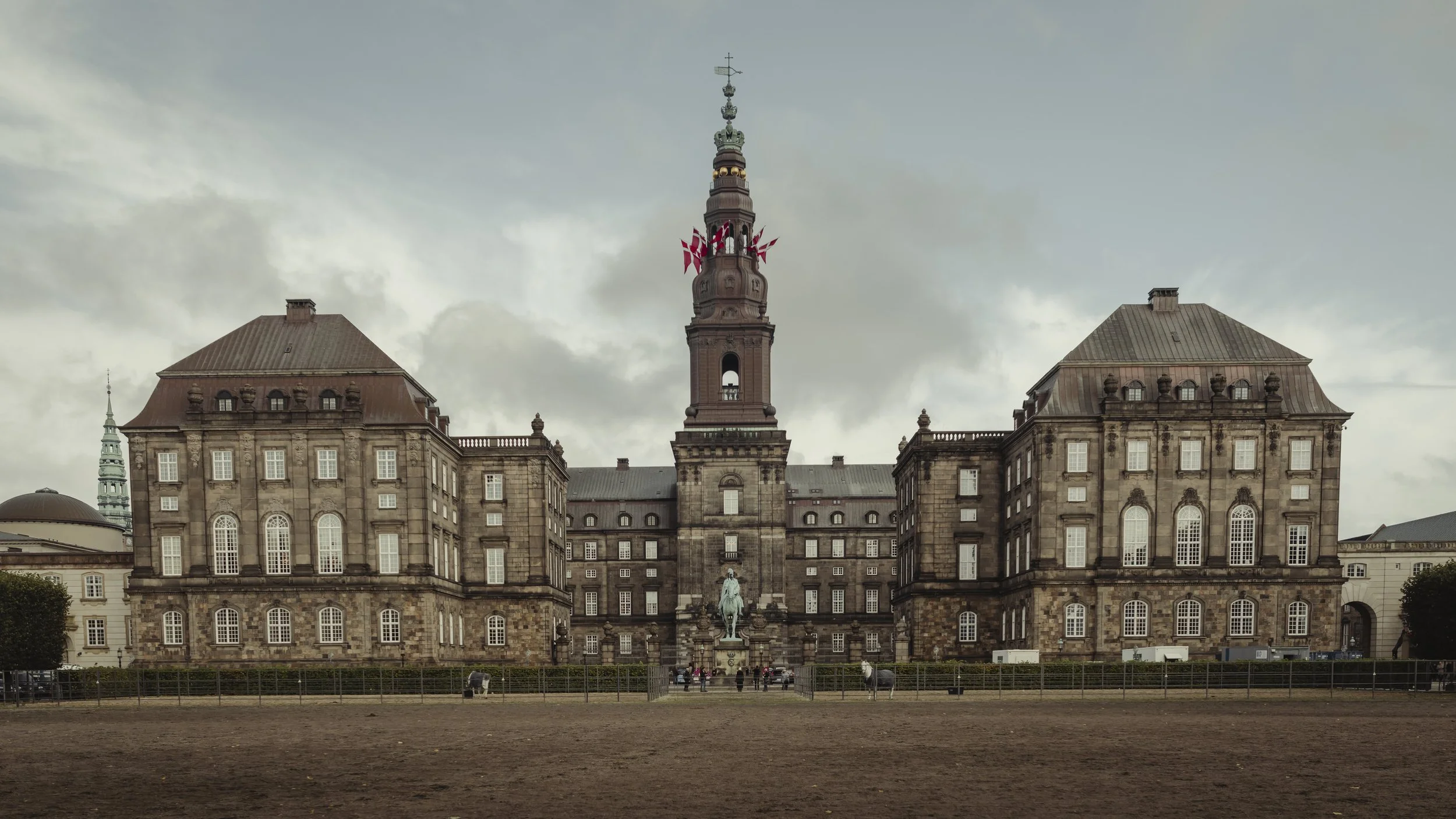 Historic government building with a central tower, statues and flags, located behind a fenced area with a dirt ground, under cloudy sky.