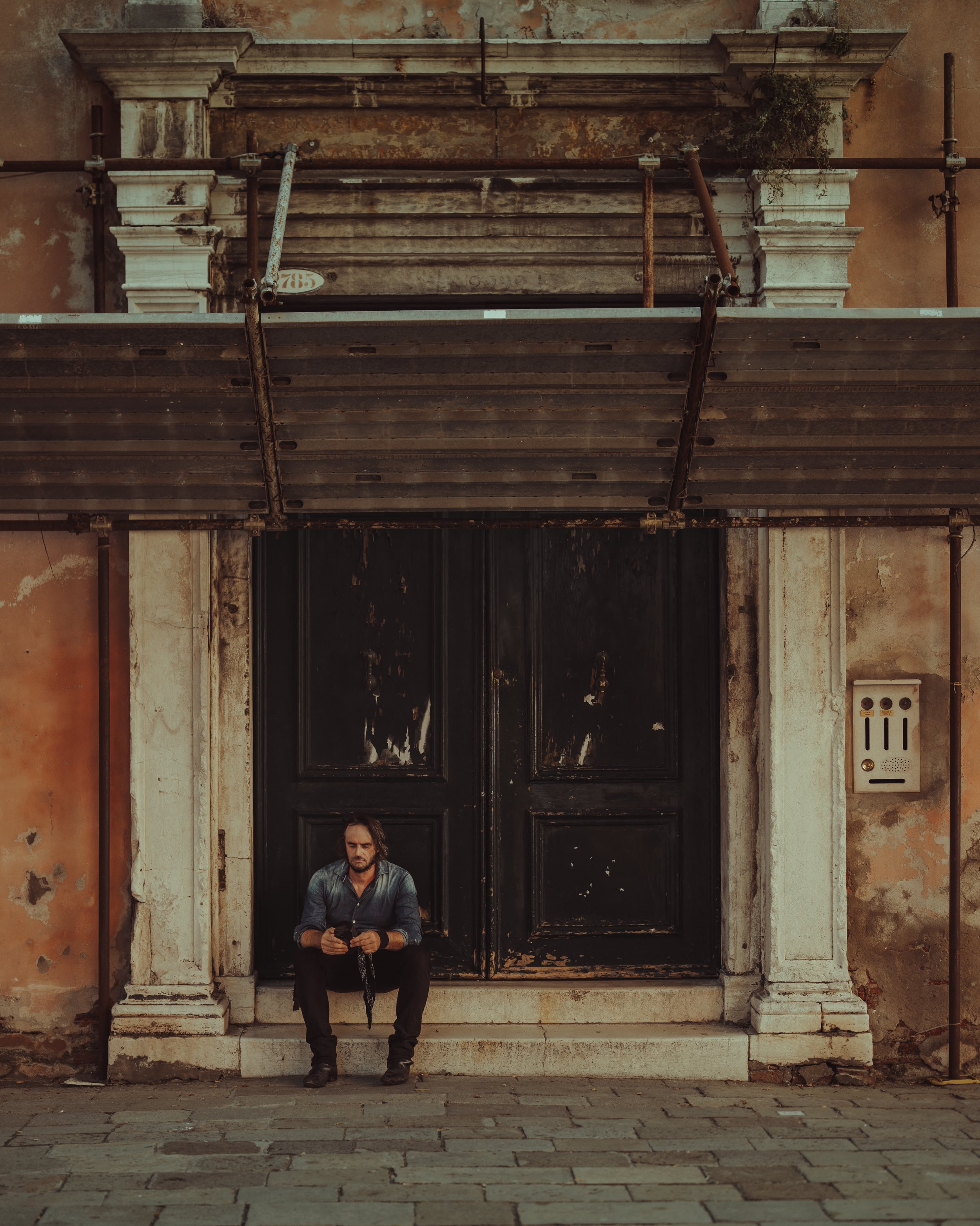 A man sitting on steps in front of a large black door with weathered paint, surrounded by peeling walls, scaffolding over the entryway, and an electrical box on the wall.