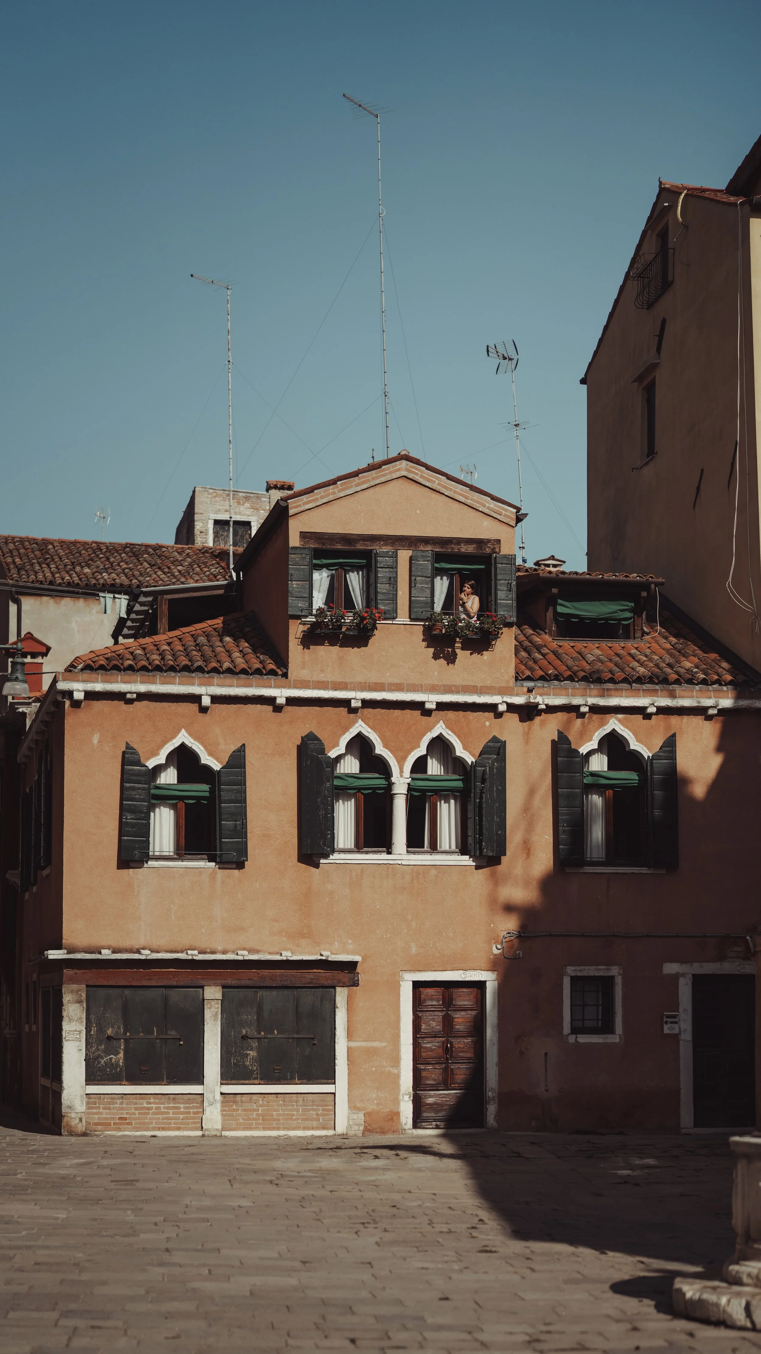 A Venetian-style apartment building with peach-colored walls, black shutters, and decorative white window arches. The top window has a person looking out, with flower boxes on the windowsills. The building has a red tile roof and is surrounded by oth