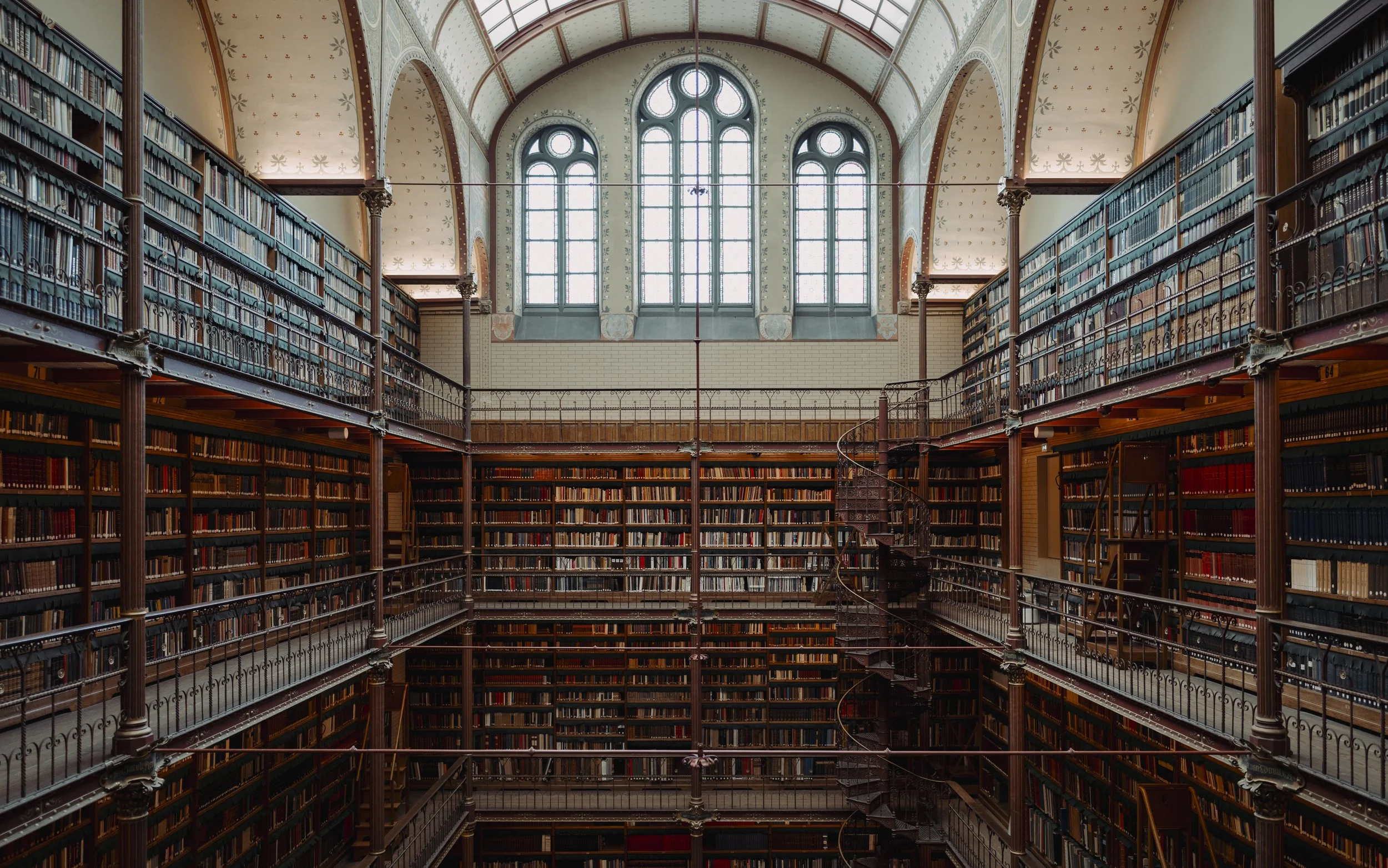 Interior of a historic library with multiple levels of bookshelves filled with books, large arched windows at the top, and a spiral staircase in the center.