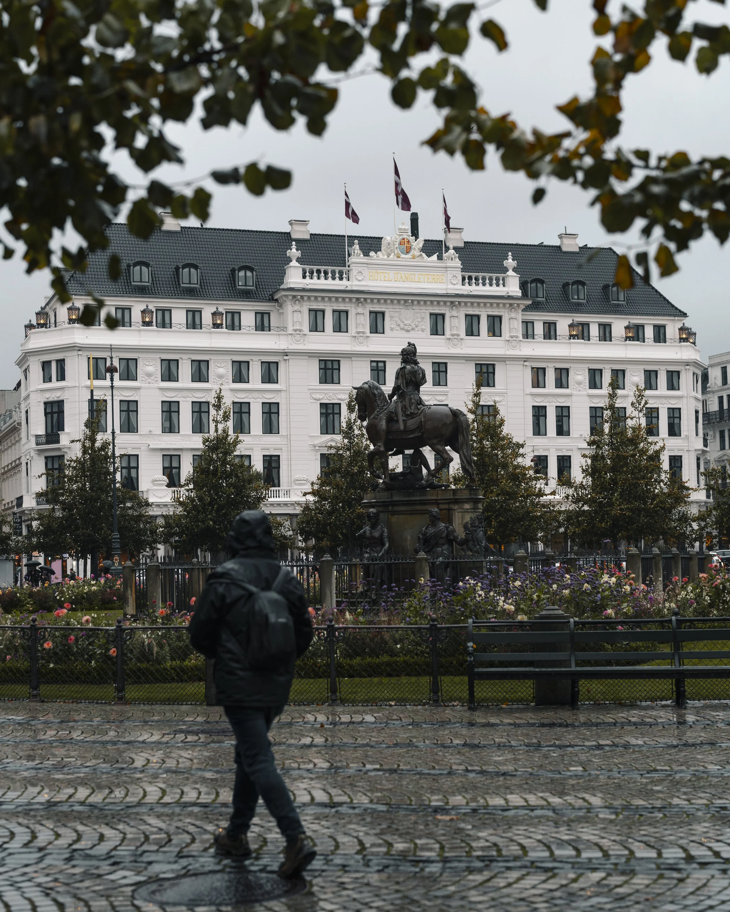 A person walking on a cobblestone street in front of a park with a bronze statue of a man on a horse, surrounded by trees, with a white grand hotel building behind and cloudy sky overhead.