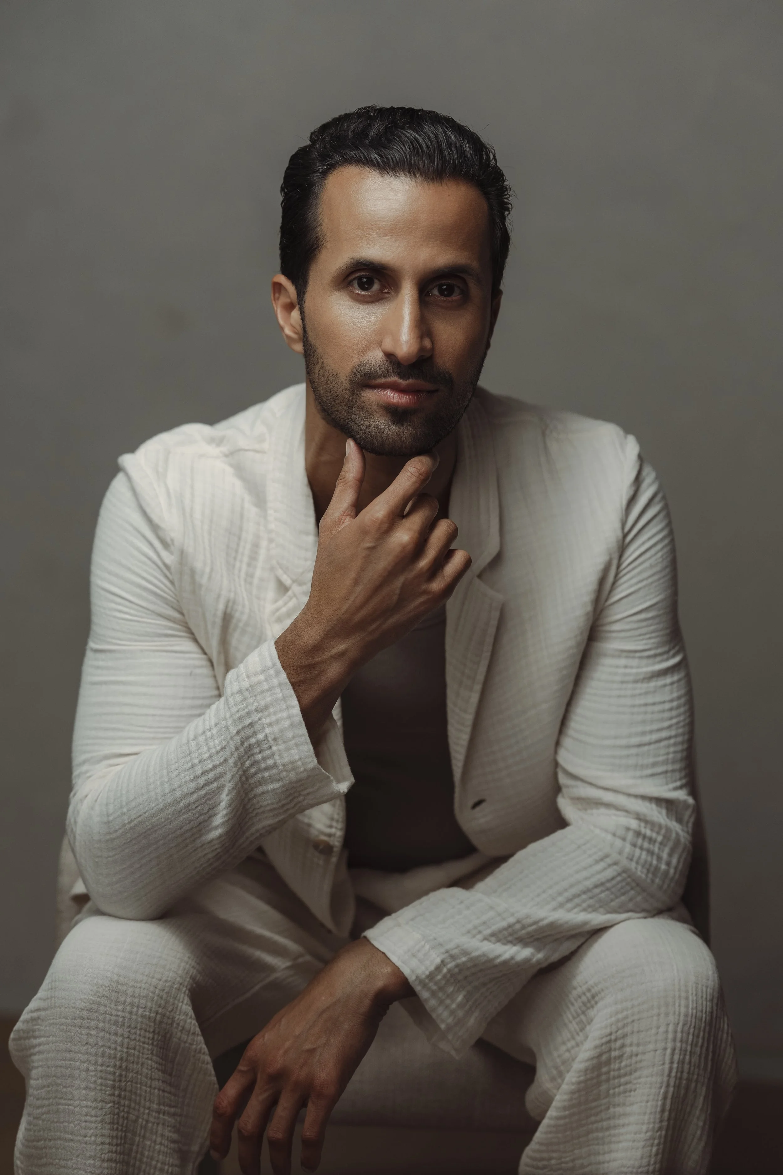 A man with dark hair and a beard wearing a cream-colored textured suit, sitting indoors against a plain gray background, with one hand on his chin and looking directly at the camera.